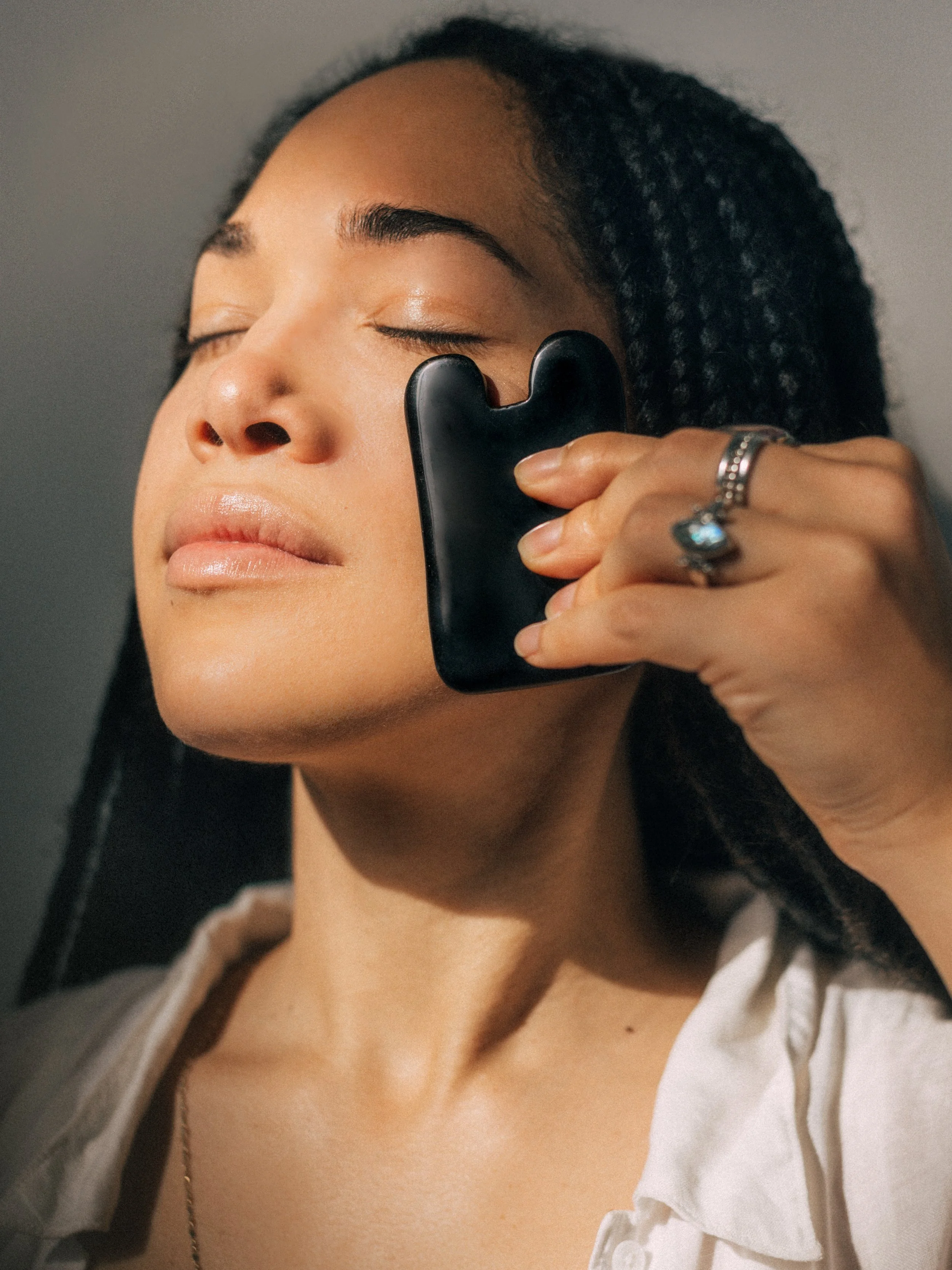 Person using a black gua sha tool on their face, eyes closed, wearing a white shirt.
