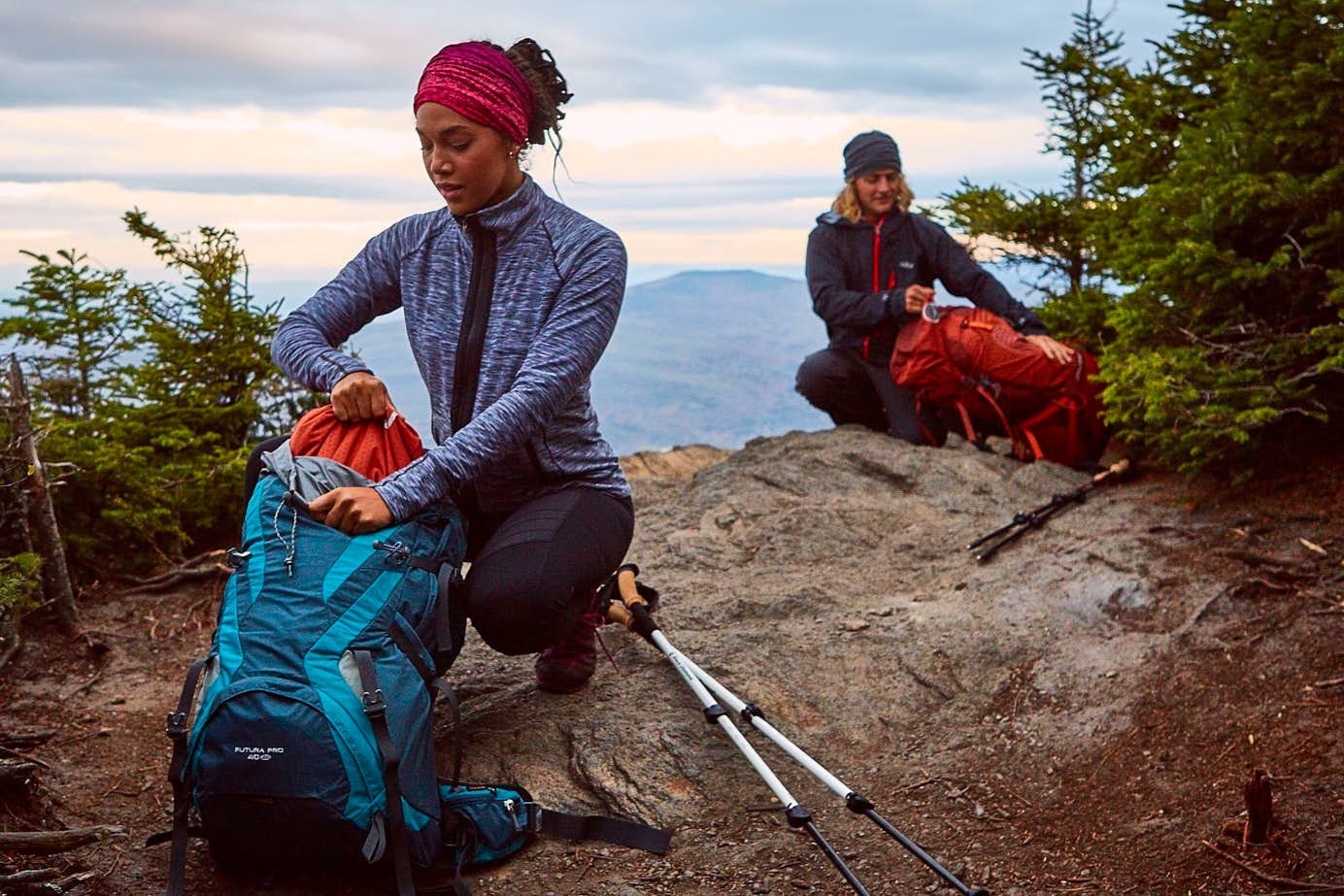 Two hikers with backpacks and trekking poles resting on a rocky mountain trail.