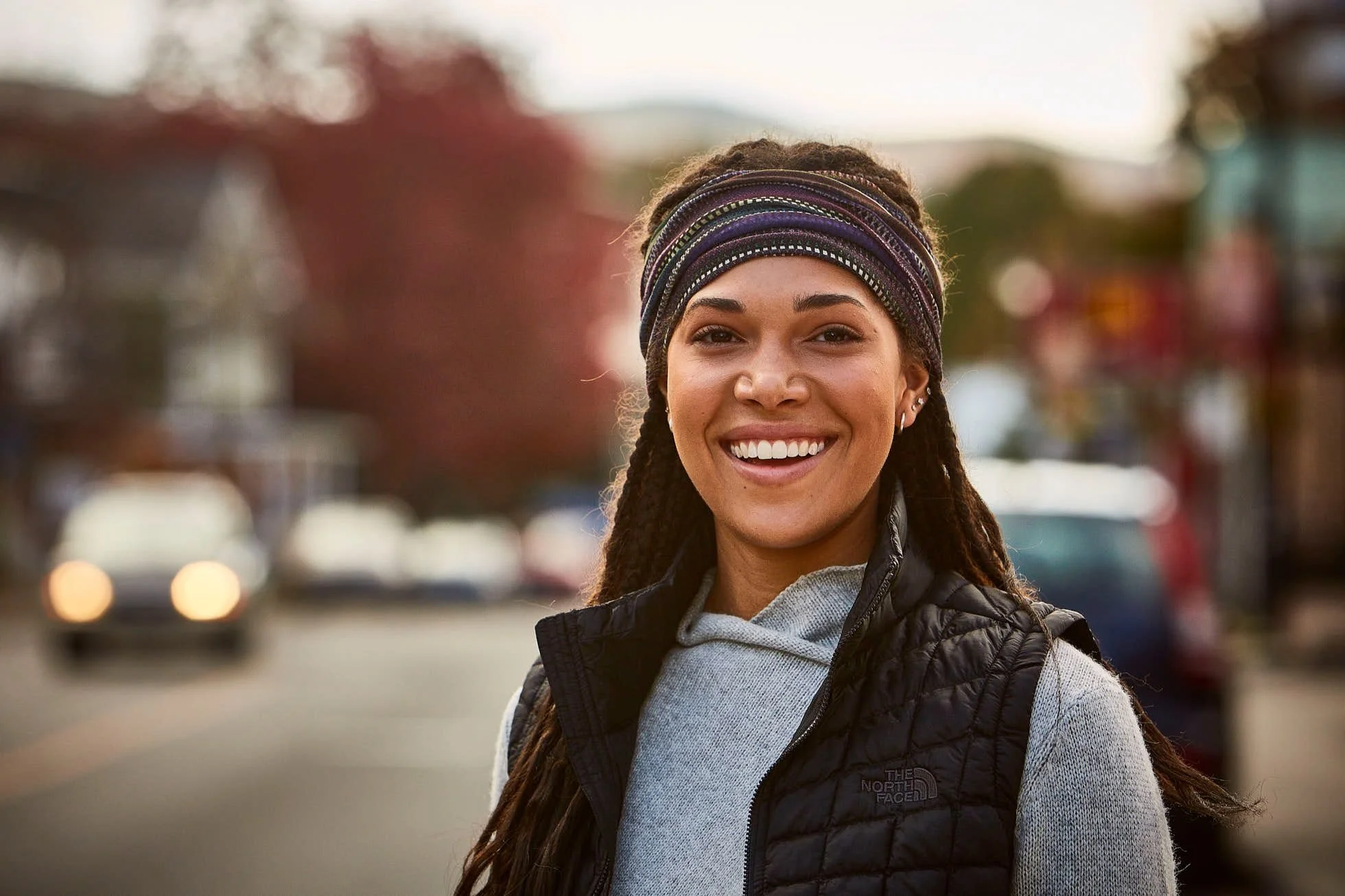 Smiling person wearing a black vest and headband outdoors on a street with a blurred background.