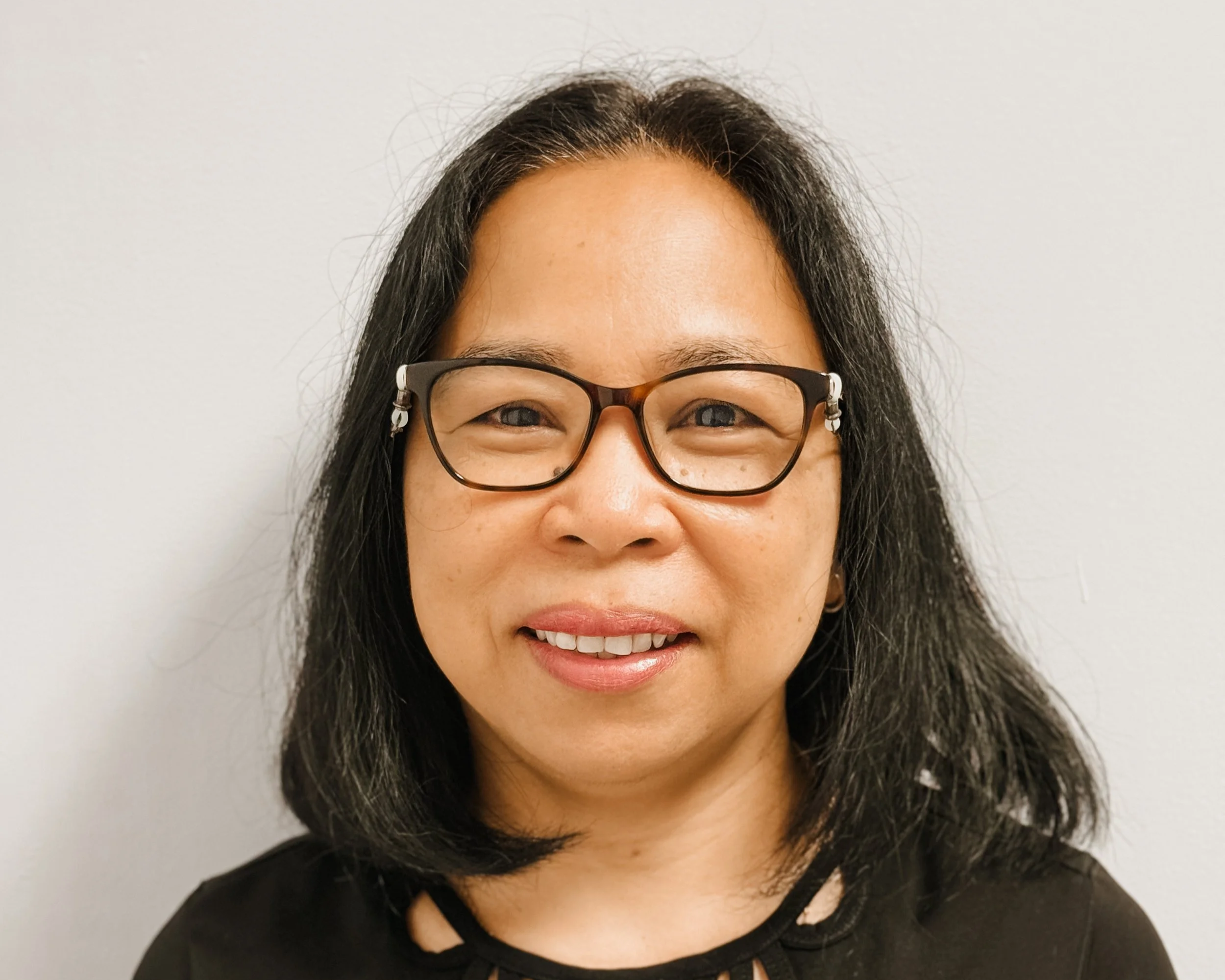 A woman with glasses and shoulder-length dark hair smiling, wearing a black top with a decorative cut-out pattern around the neckline, standing against a plain white wall.