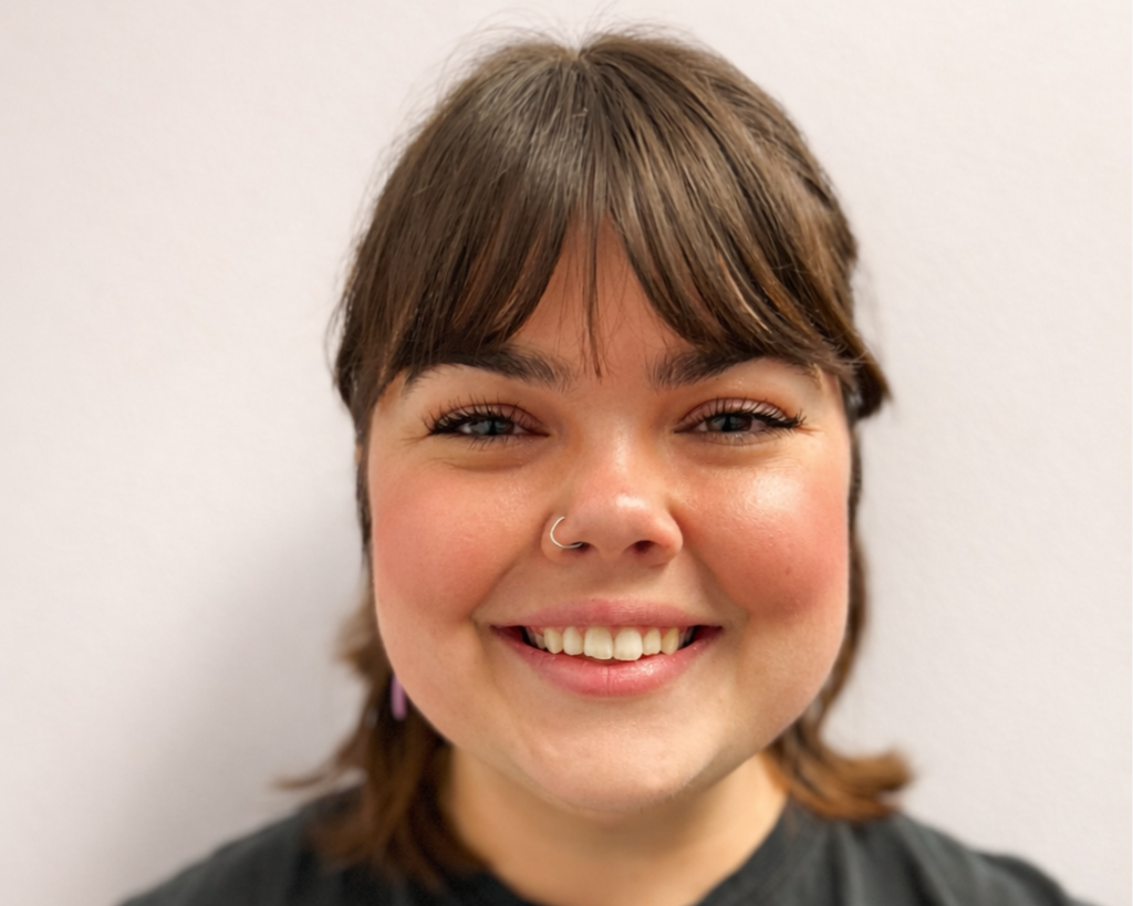 A young woman with shoulder-length brown hair, wearing a nose ring and a black T-shirt, smiling against a plain white background.