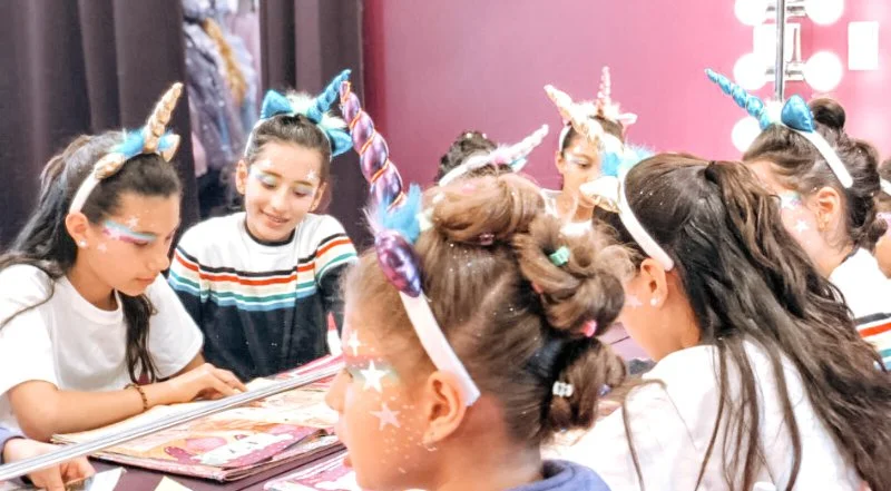 Kids dressed as unicorns with cone-shaped horns, colorful wigs, and star and glitter face paint, gathered around a table reading books in a decorated room.