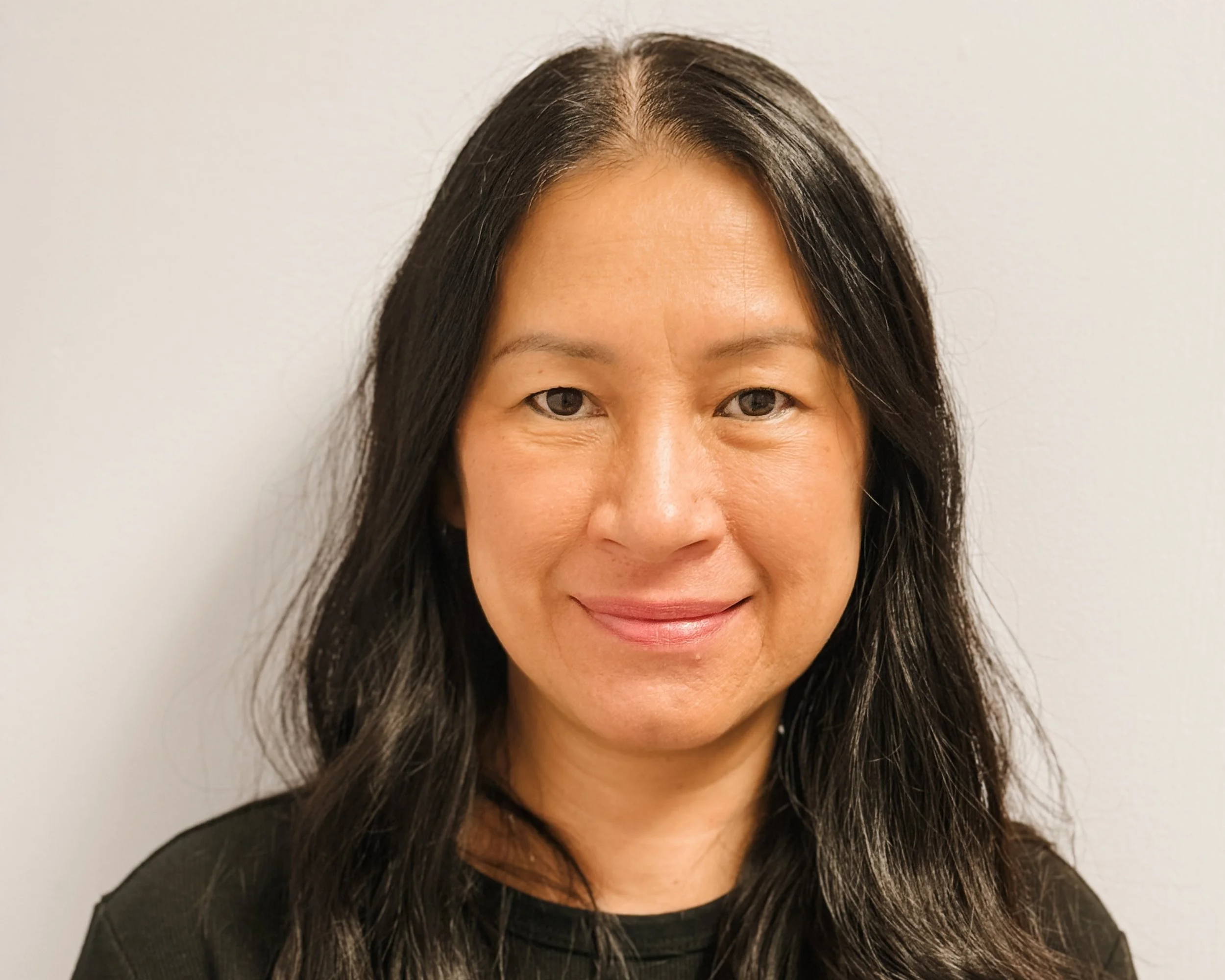 A woman with long black hair and a slight smile, wearing a black top, stands against a plain off-white wall.