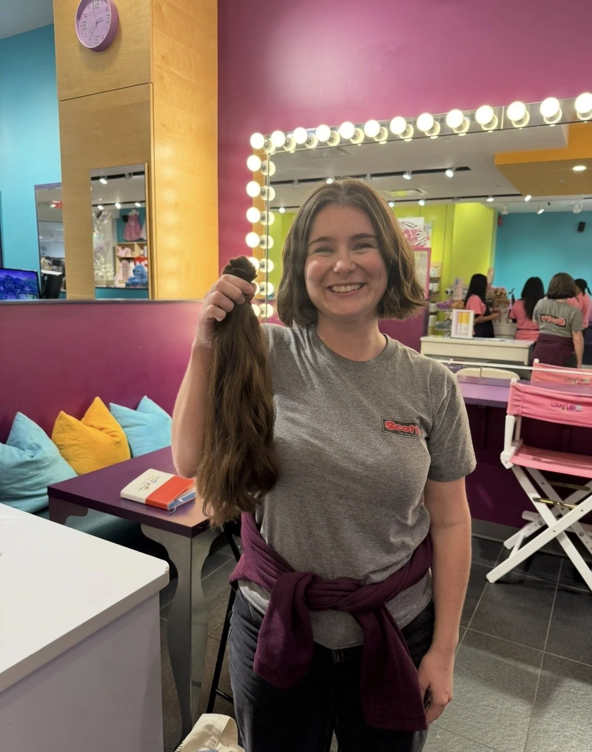 A smiling woman in a gray t-shirt holding a long strand of hair inside a colorful salon with bright lights, pink, blue, and yellow walls, and a mirror with bulbs. Other people are working in the background.