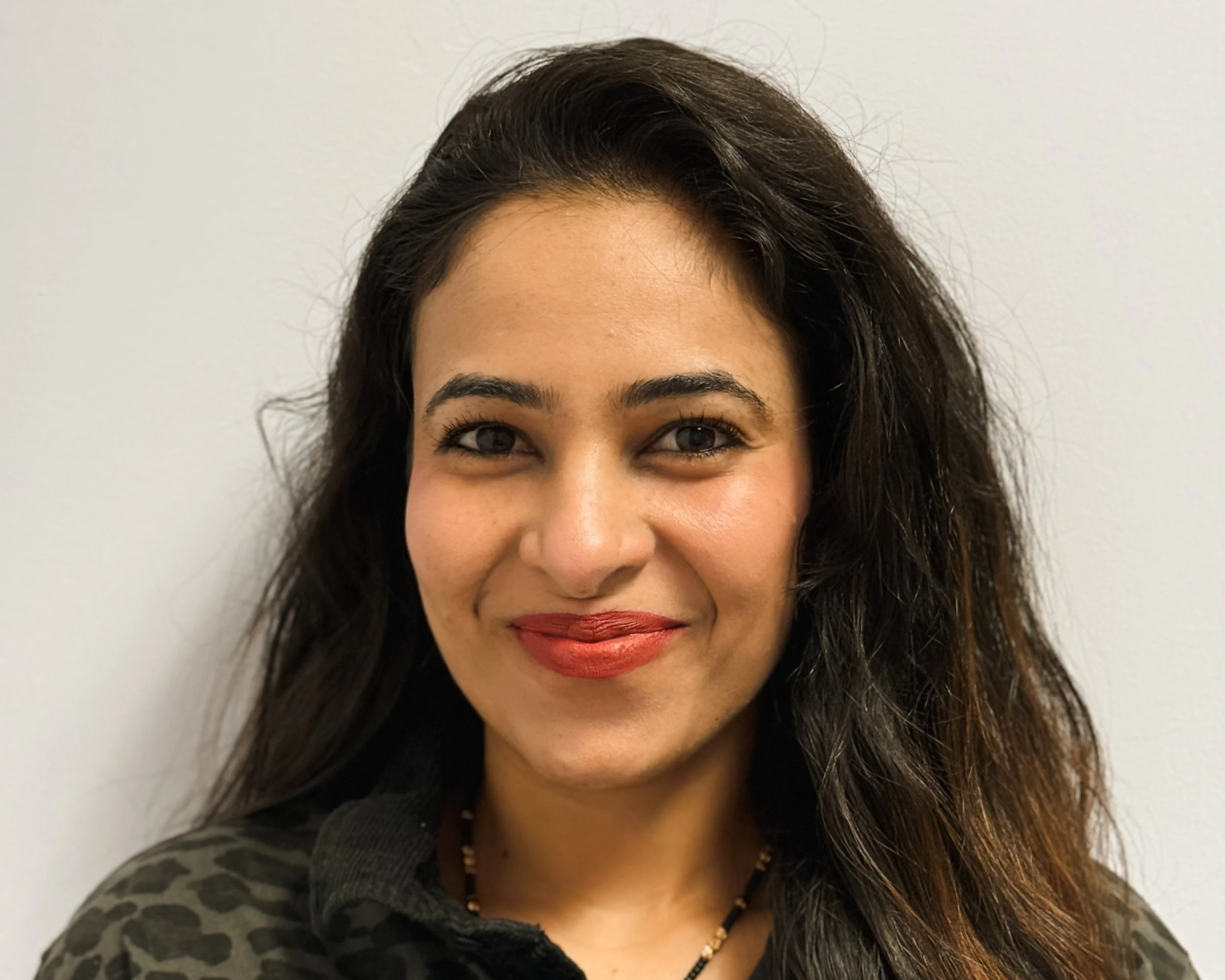 A woman with long, dark wavy hair and medium skin tone, smiling slightly, wearing a black shirt with a leopard print pattern and a beaded necklace, standing against a plain white wall.
