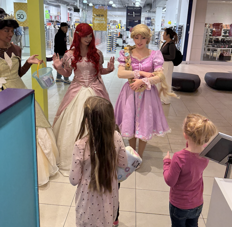 Two women dressed as princesses, one with red hair and the other with blonde hair, interacting with two young girls in a shopping mall.