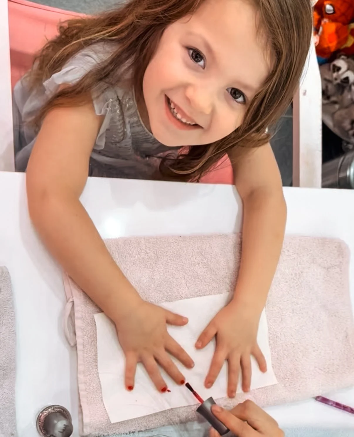 A young girl smiling, sitting at a table with her hands placed on a nail salon table, getting her nails painted red by a person in front of her.
