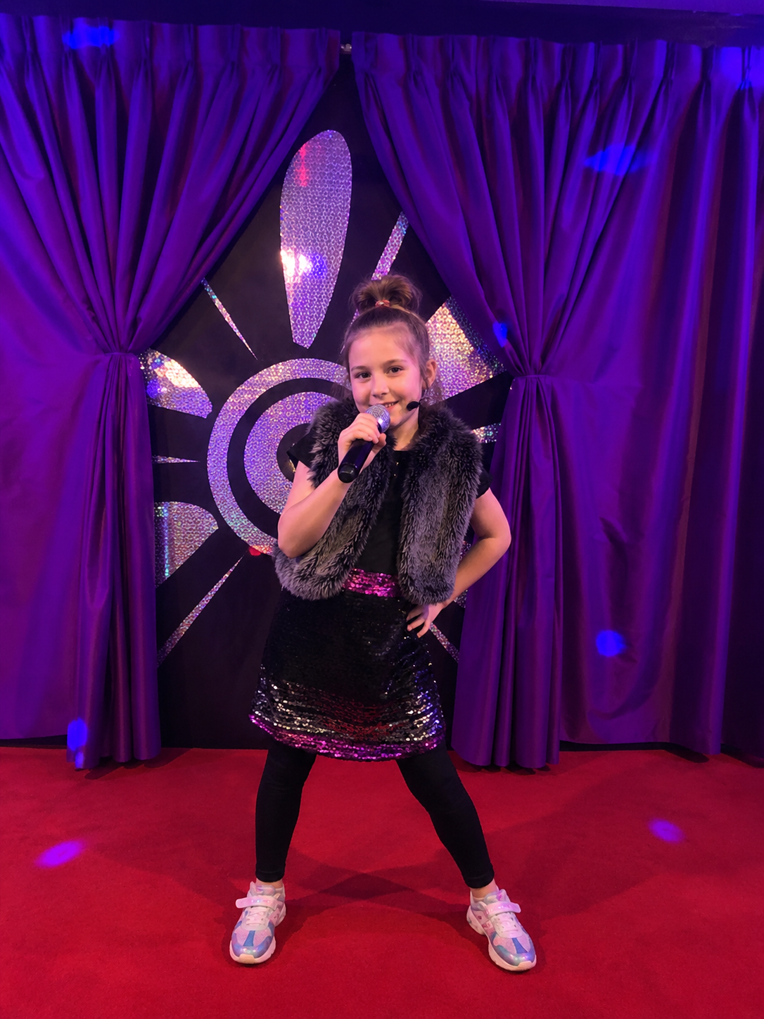 A young girl is standing on a red carpet stage with purple curtains, holding a microphone and smiling. Behind her is a black backdrop with a colorful, glittery geometric design. She is wearing a black and purple outfit with a faux fur vest and sneakers.