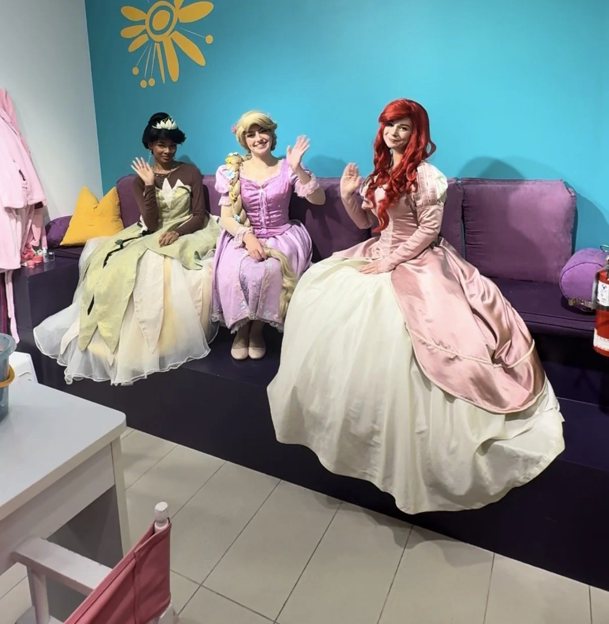 Three women dressed as Disney princesses sitting on a purple couch, waving at the camera in a colorful room with a blue wall and floral decoration.