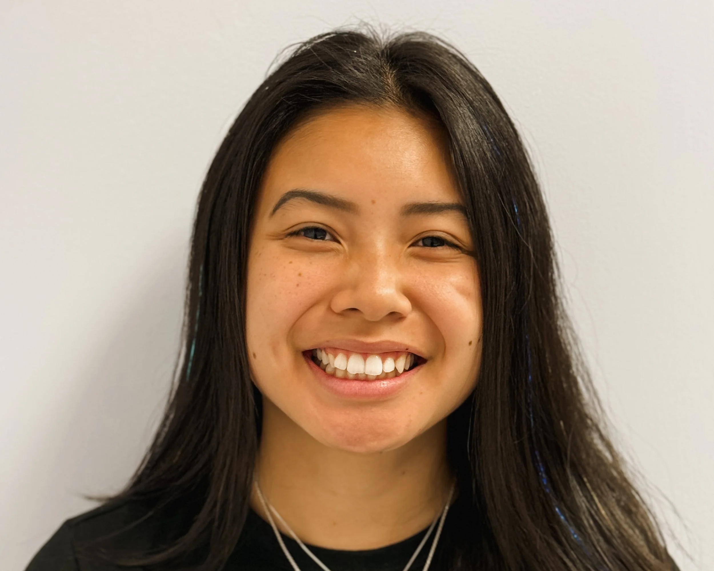 Close-up of a young woman with long dark hair, smiling, wearing layered necklaces and a black shirt, against a plain light-colored wall.