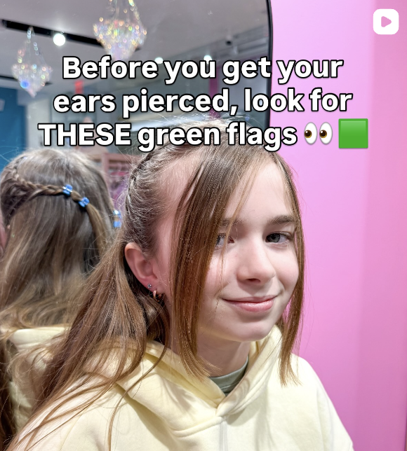 A girl with light brown hair pulled back, wearing a cream hoodie, smiling slightly, standing in front of a pink wall. There is a reflection of another girl with blonde hair and a blue hair clip in the mirror behind her. The image has overlay text that talks about looking for green flags before getting ears pierced.