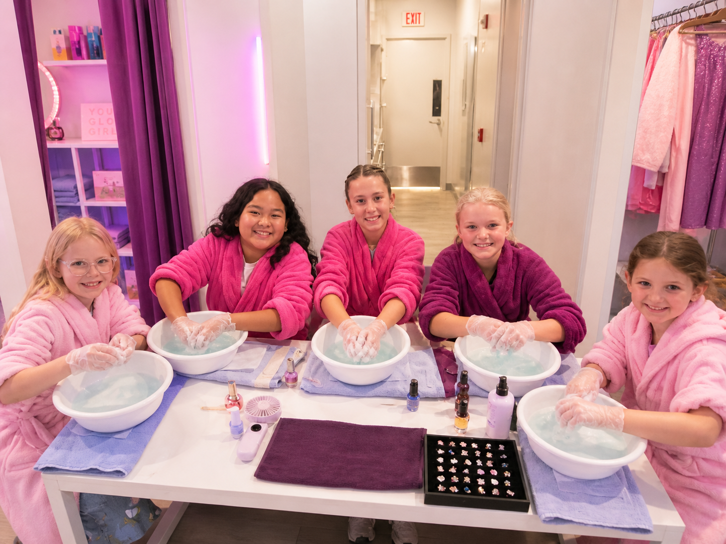 Five young girls wearing pink and purple robes sitting at a table with bowls of water and nail polish bottles, practicing manicures in a spa or nail salon.