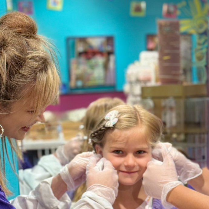 A young girl getting her ears pierced at a jewelry store with a woman, likely a staff member, assisting her. The girl has braided hair and is wearing a white shirt, smiling as the piercing is done. The background shows colorful decorations and shelves with products.