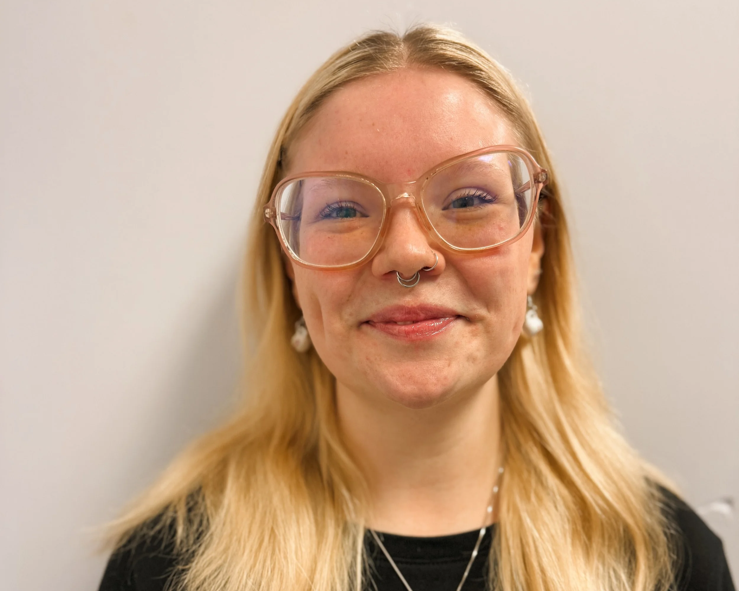 Close-up of a smiling woman with blonde hair, wearing large pink glasses, a septum piercing, star-shaped earrings, a star necklace, and a black top, standing against a plain light-colored wall.