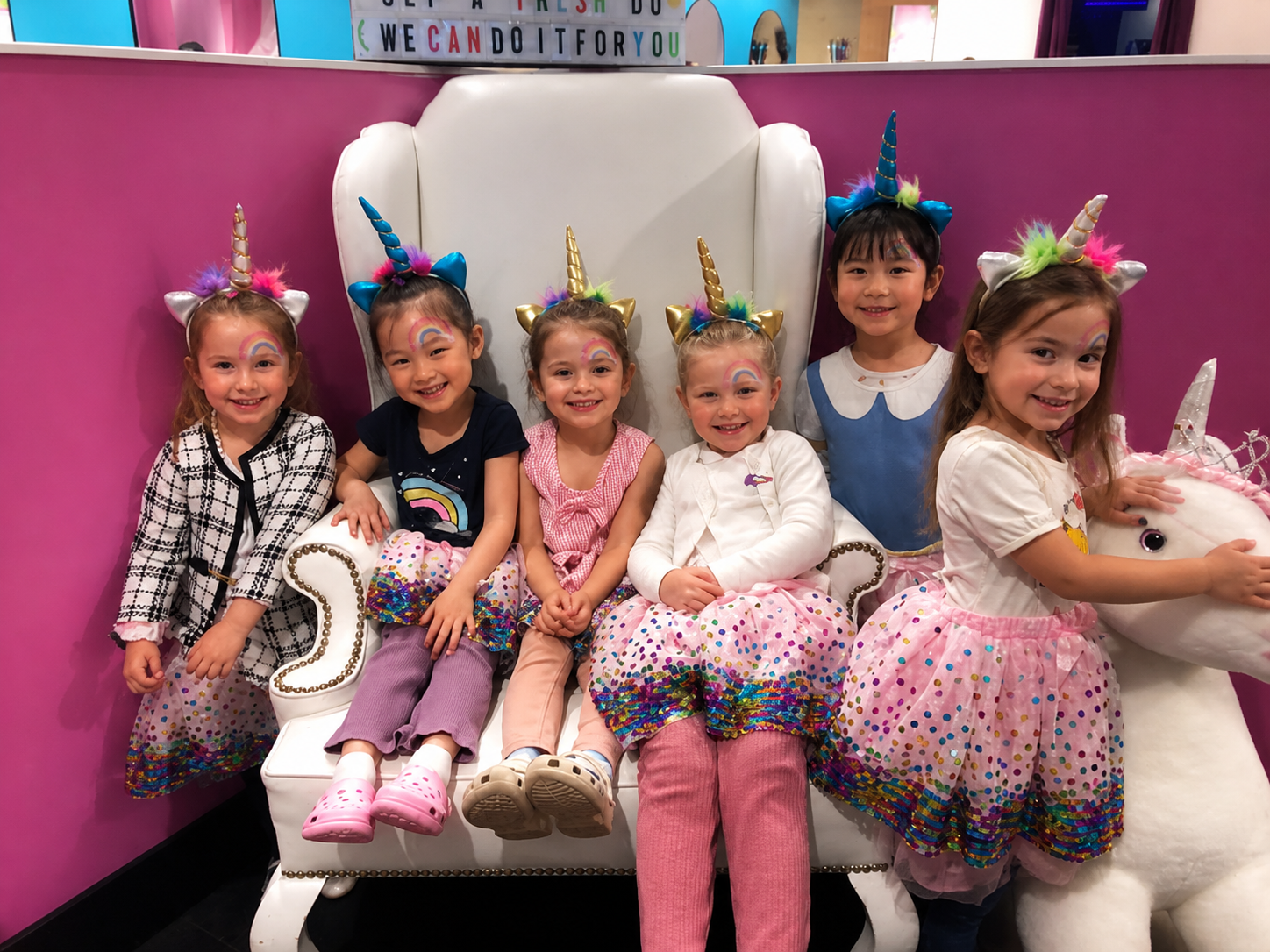 Six young girls dressed as unicorns sitting on a white, heart-shaped couch with pink walls in the background.