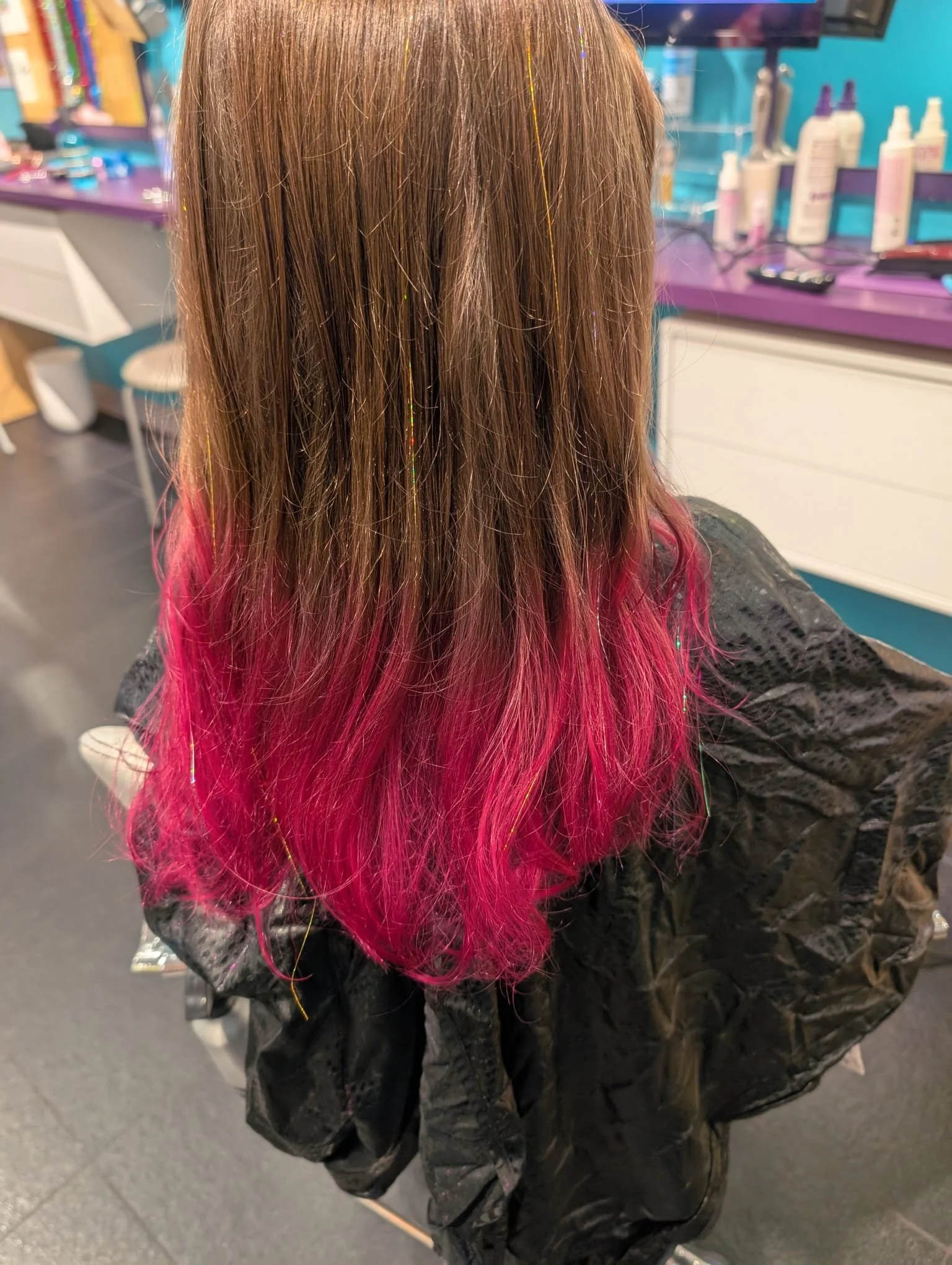 Person with long brown hair that transitions to pink at the ends, sitting in a salon chair, wearing a black cape, with salon tools and hair products visible in the background.
