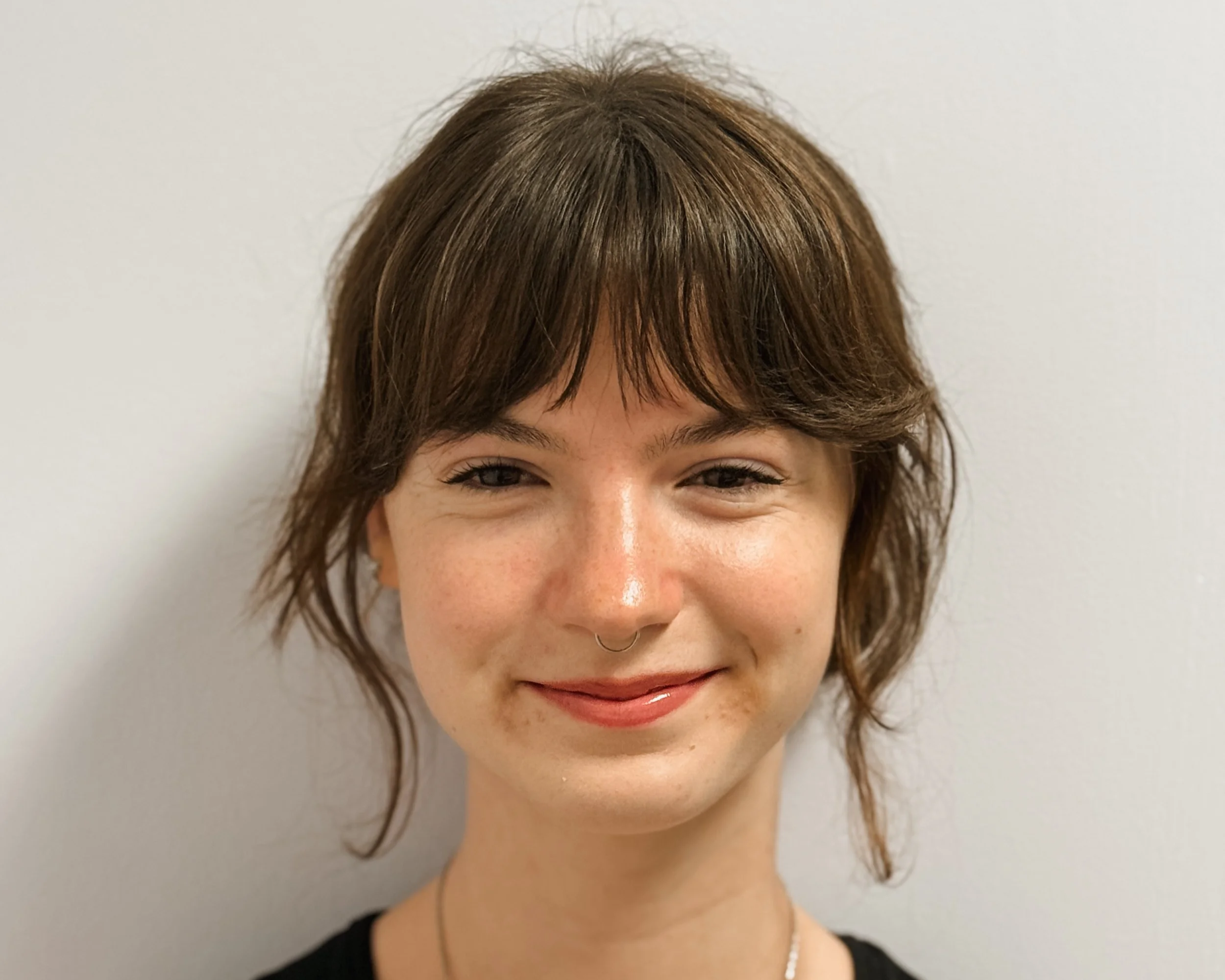 Close-up of a smiling woman with short brown hair, wearing a black sleeveless top and a silver necklace, standing against a plain light background.