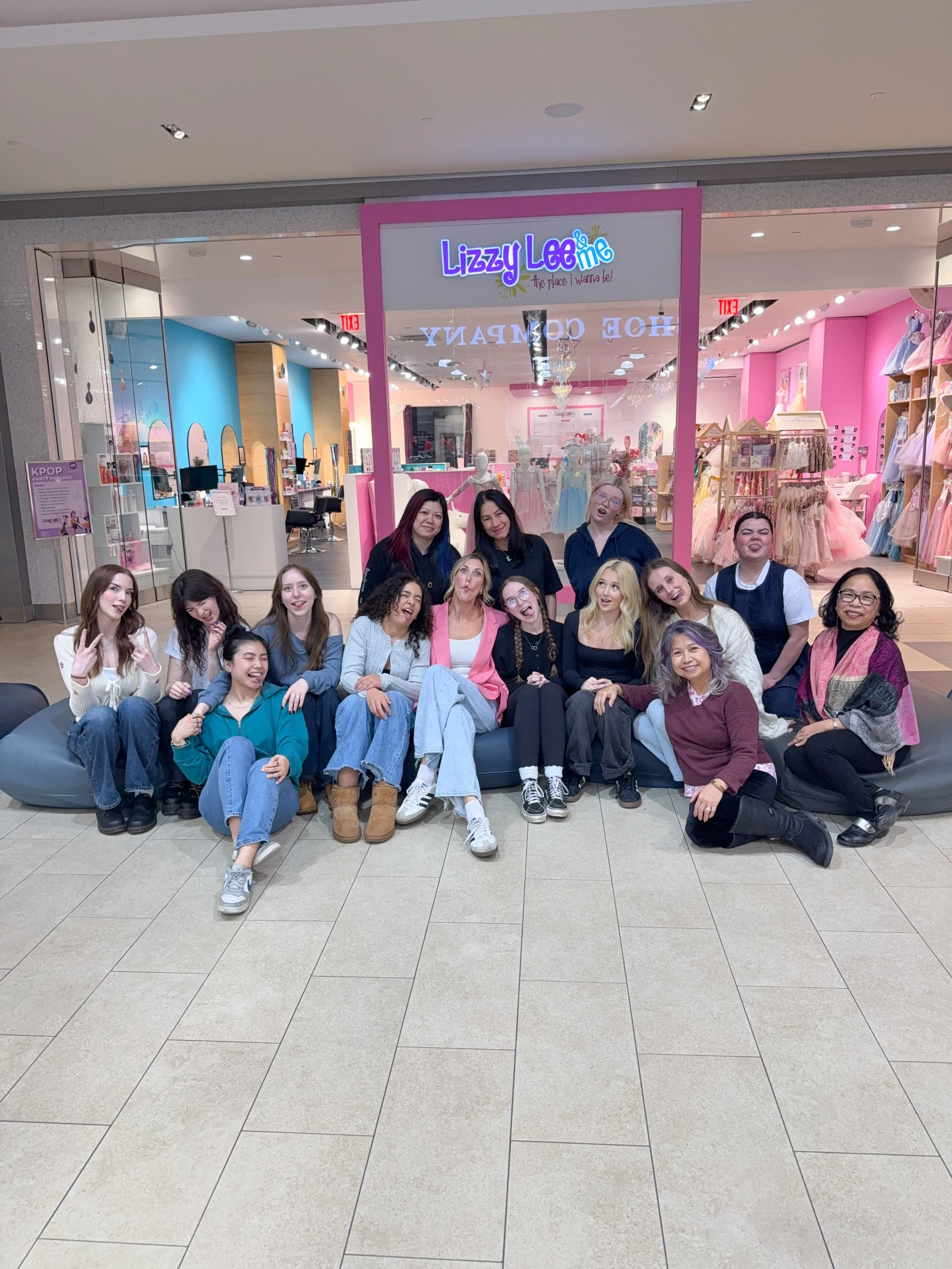 Group of people sitting on a black couch in front of a store called Lizzy Lee in a shopping mall. The store displays ballerina dresses and has a pink and white color theme.