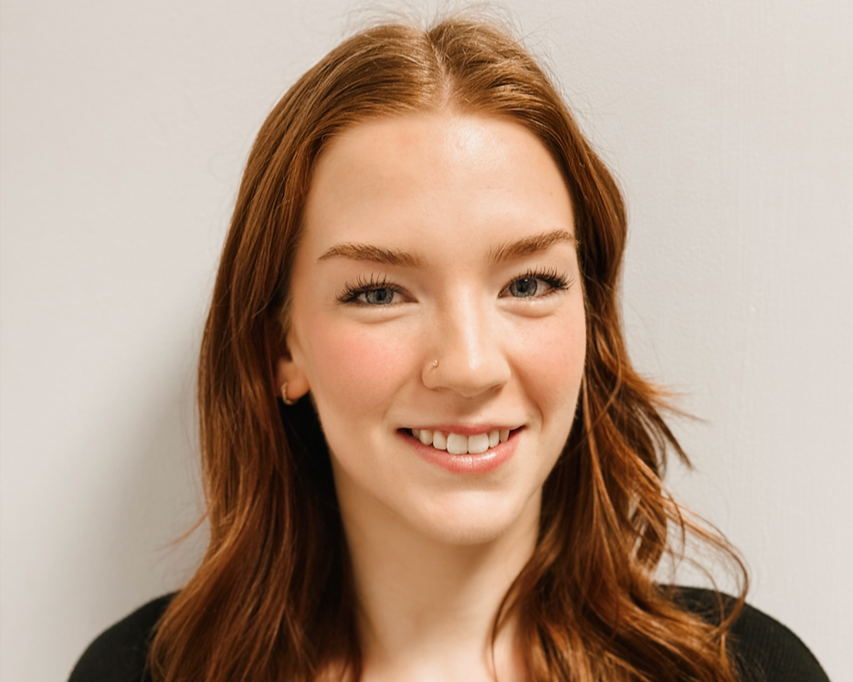 Close-up of a young woman with long red hair, blue eyes, and a nose piercing, smiling at the camera, wearing a black top.