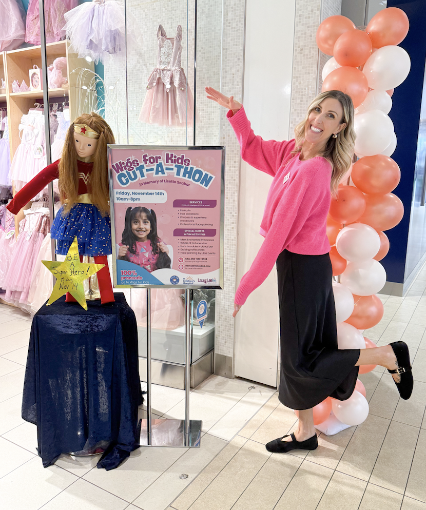A woman in a pink sweater and black skirt poses playfully next to a colorful balloon column inside a shopping mall. She is smiling and pointing at a sign for a kids' charity event called Wig for Kids CUT-A-THON, with a display of a doll dressed as Wo
