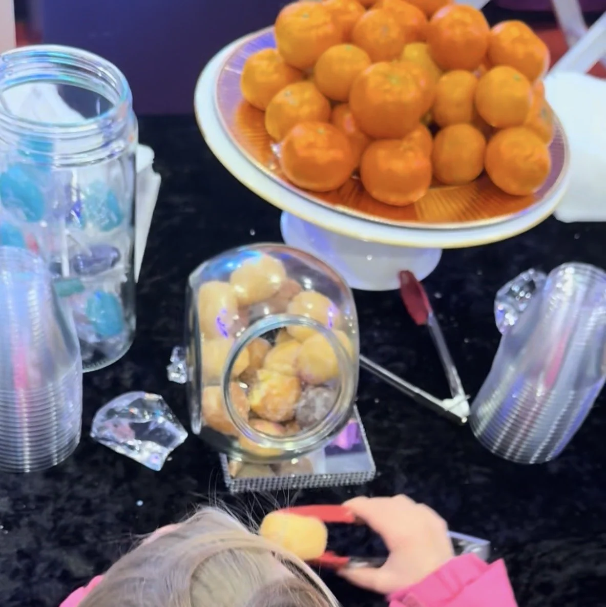 A table with a large plate of bright orange tangerines, multiple empty clear plastic cups stacked, and a person with gray hair and pink sleeves scooping a tangerine with red tongs.