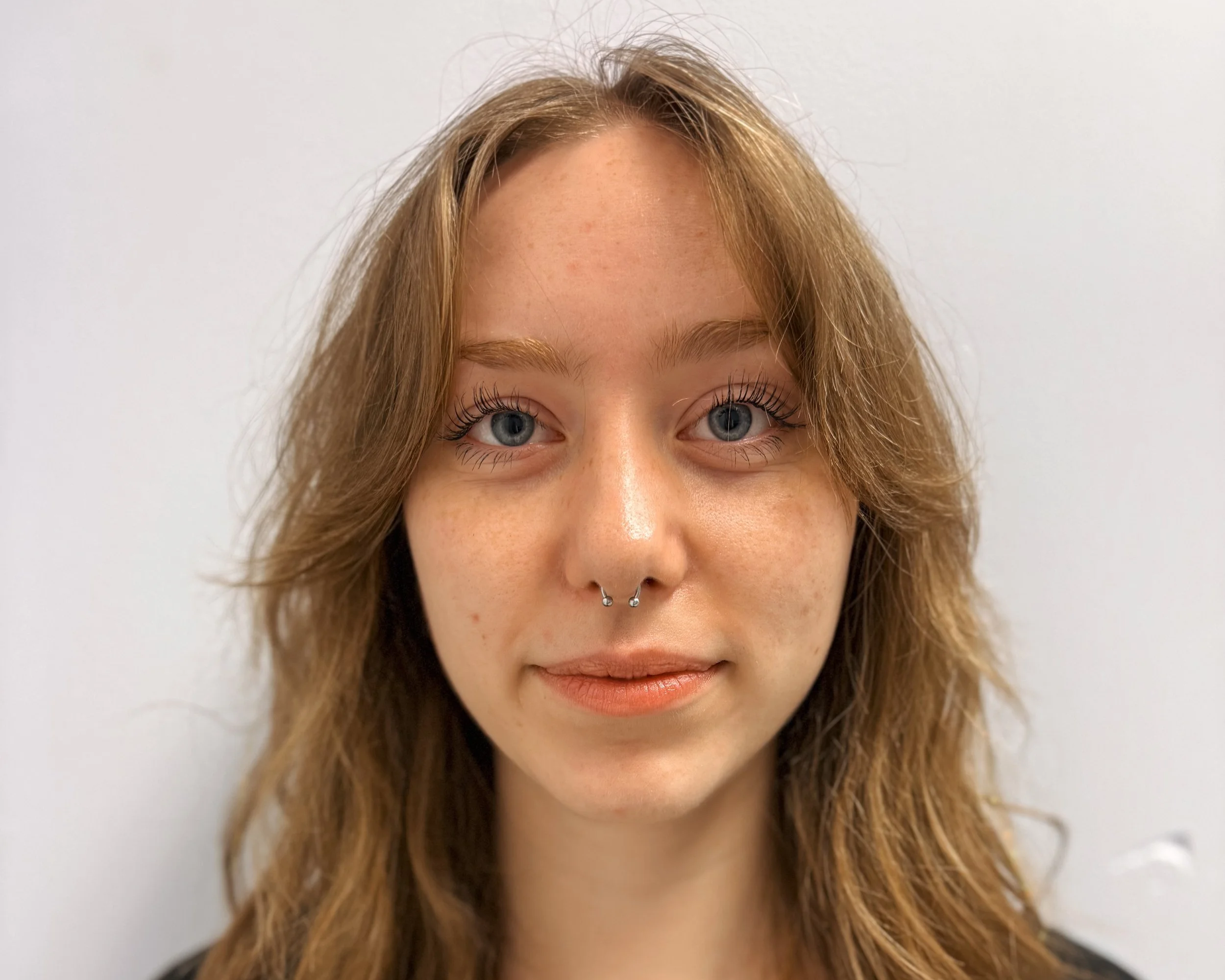 A young woman with long, wavy red hair and blue eyes is looking at the camera with a slight smile. She has a septum piercing and is wearing a black top with a gold necklace. The background is plain and white.