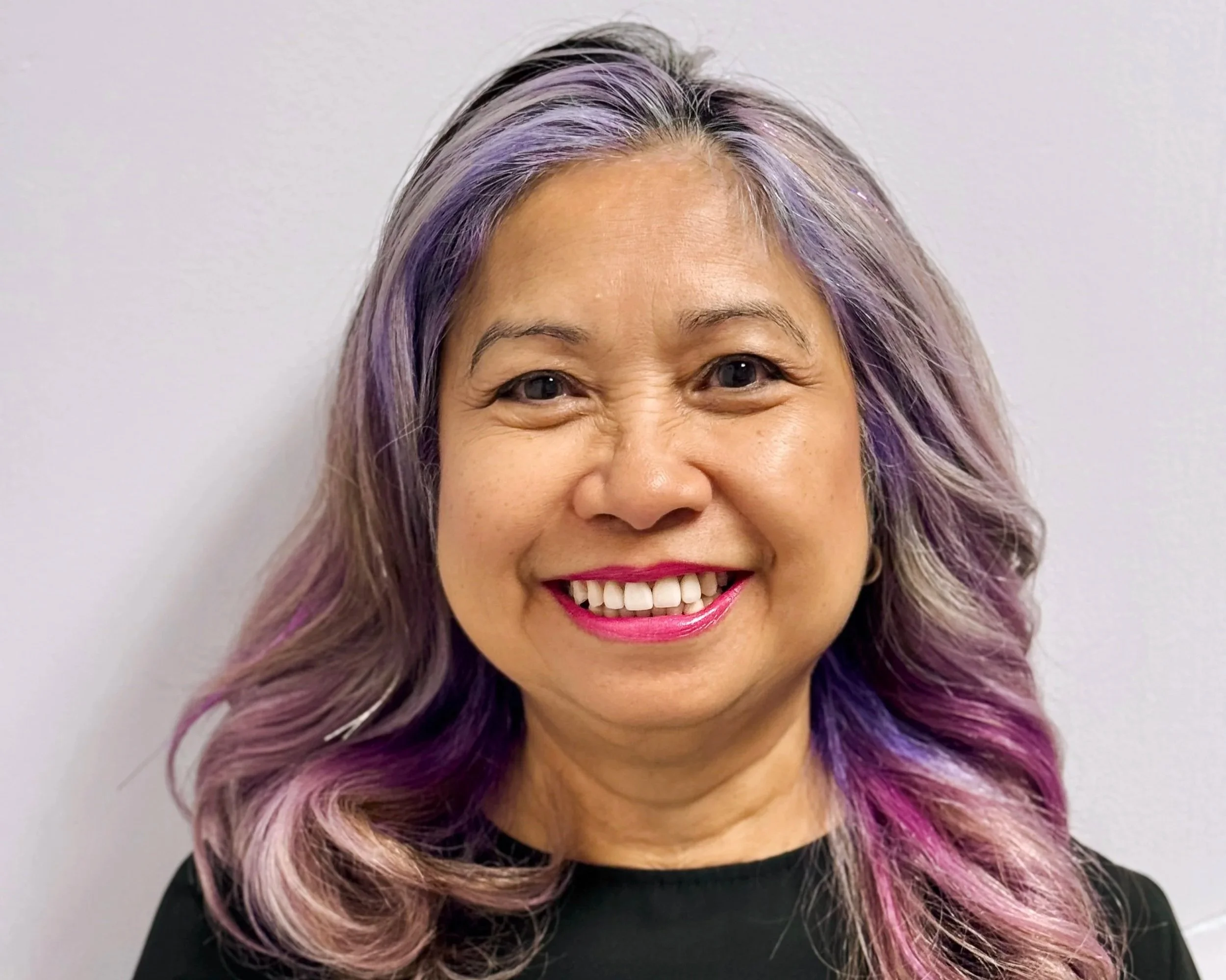 Close-up of a woman with shoulder-length hair dyed in shades of purple and pink, smiling against a plain white wall.