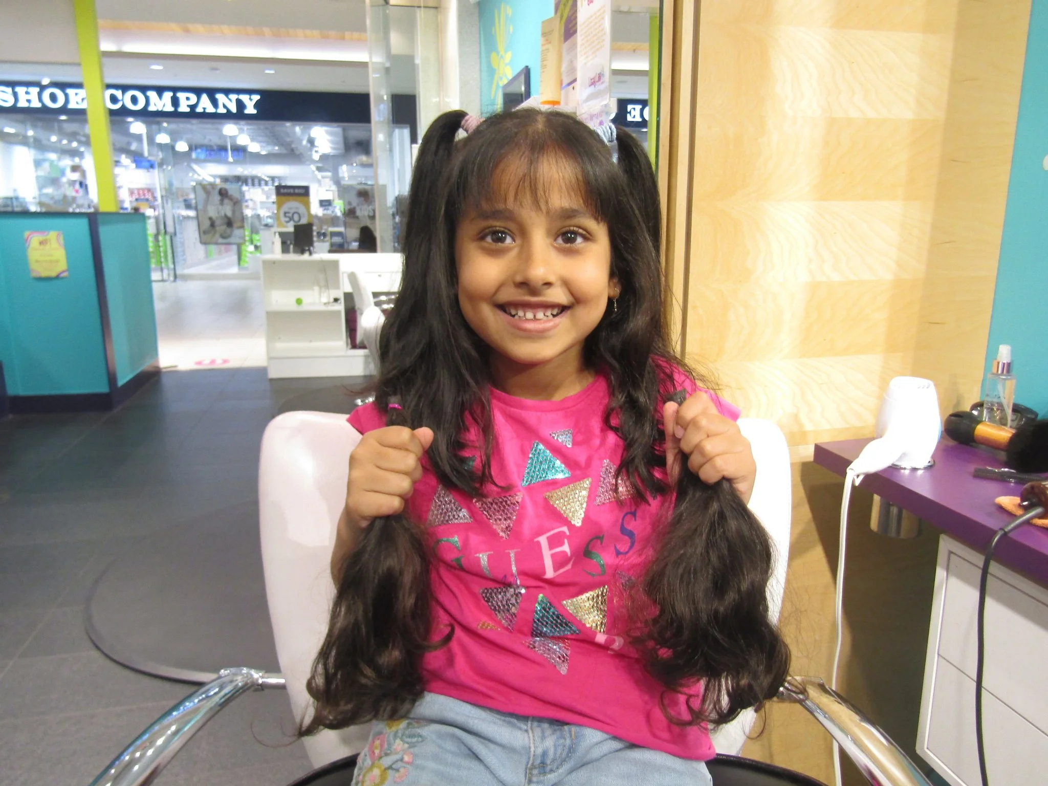 A young girl with long dark hair smiling and holding her fists up inside a shopping mall or store, with a shoe store sign visible in the background.