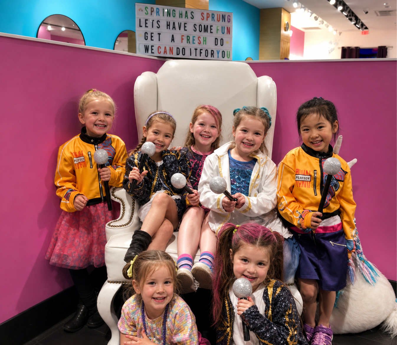 Group of young girls sitting on a large white chair in a colorful indoor setting, holding silver microphones, smiling for the camera, with a colorful sign in the background that says "Spring Has Sprung, Let's Have Some Fun, Get a Fresh Do."