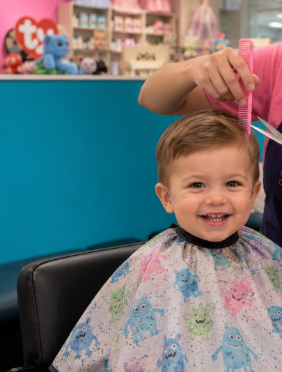 A young boy sitting in a barber's chair, smiling as a barber cuts his hair. The boy is wearing a cape with colorful monster prints, and the barber is holding scissors and a pink comb.