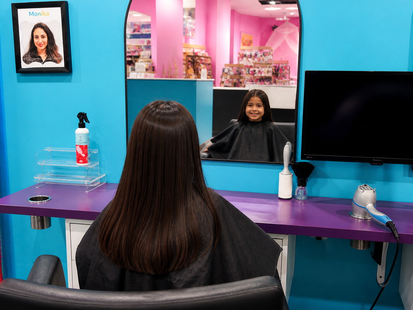 A young girl with long brown hair sitting in a salon chair, looking at herself in a mirror and smiling. The salon has bright blue and purple walls, and a framed picture of a woman with the name 'Monika' on the wall. Various hair styling tools and products are placed on the purple countertop.