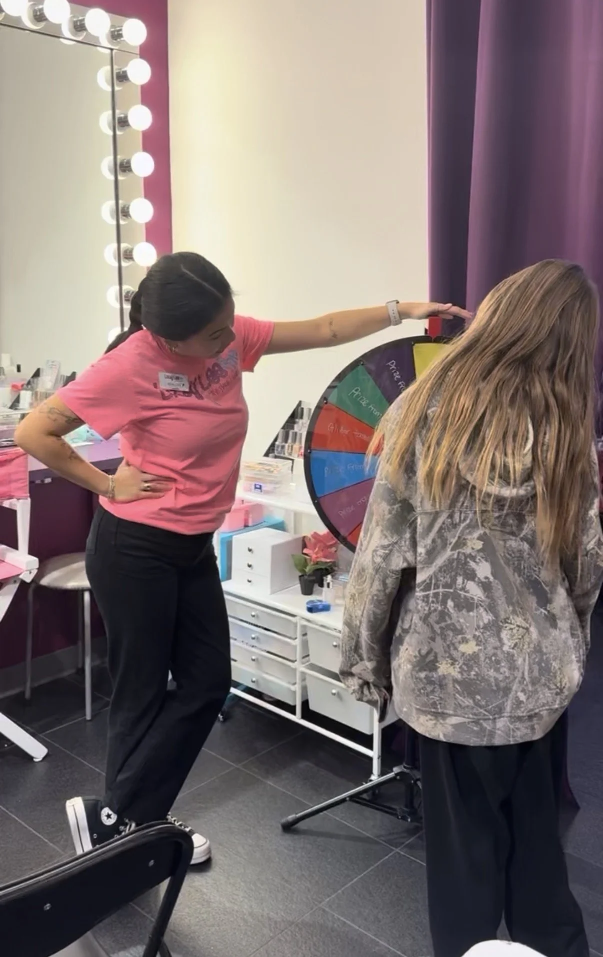 A woman in a pink shirt is orienting herself next to a spinning prize wheel with colored sections, inside a brightly lit room with a mirror and vanity lights. Another person with long blond hair and a patterned gray hoodie stands nearby, facing the w