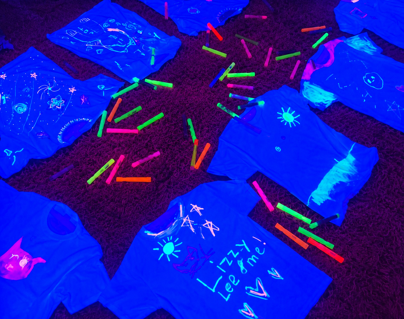 Group of glow-in-the-dark t-shirts and colorful glow sticks arranged on a carpeted floor.