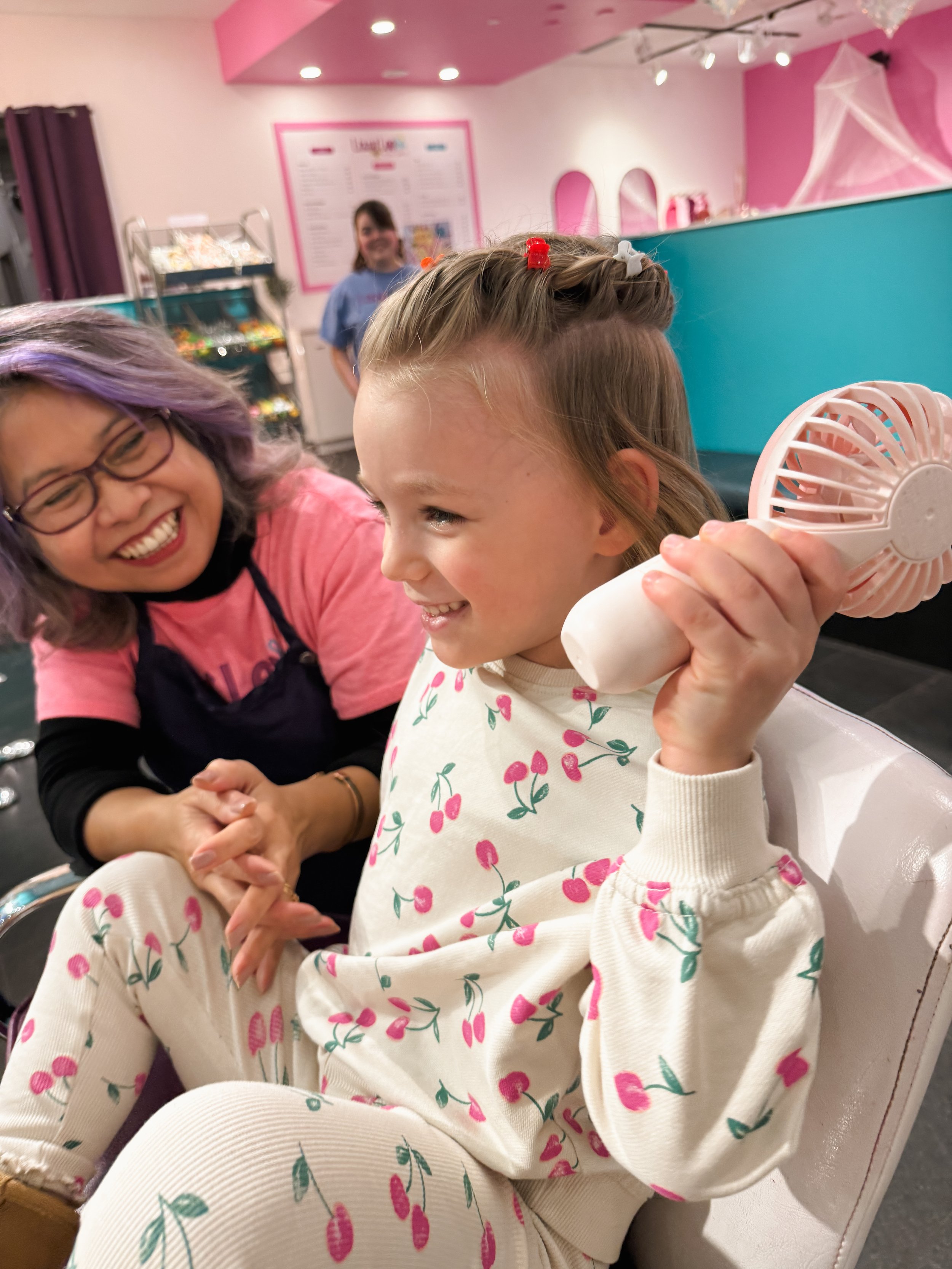 A young girl with light brown hair styled with colorful hair clips is smiling and holding a small handheld fan while sitting in a salon or beauty shop. An adult with purple hair and glasses is smiling and holding her hands, sitting nearby. A woman in a blue uniform is in the blurred background, in front of pink and blue walls decorated with hanging spiderwebs.