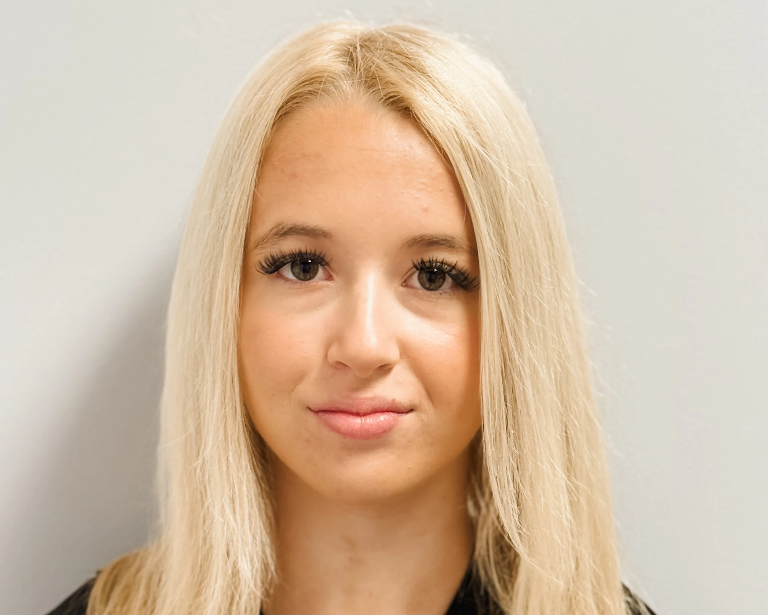 Close-up of a woman with blonde hair, hazel eyes, black top, smiling softly, against a light gray background.