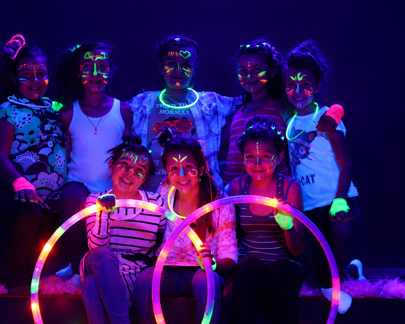 Group of children with face paint illuminated by black light, holding glow sticks and smiling.
