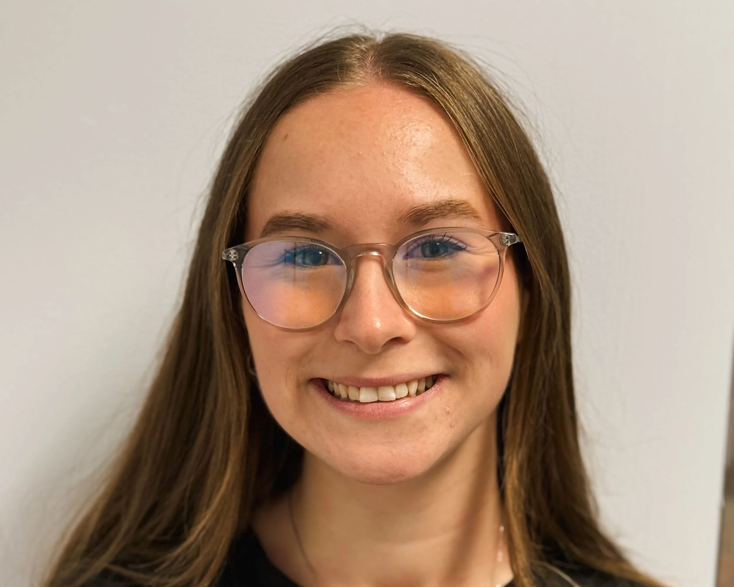 Close-up of a woman smiling, wearing glasses, a black top, and a silver necklace with three connected circles, against a plain light-colored background.
