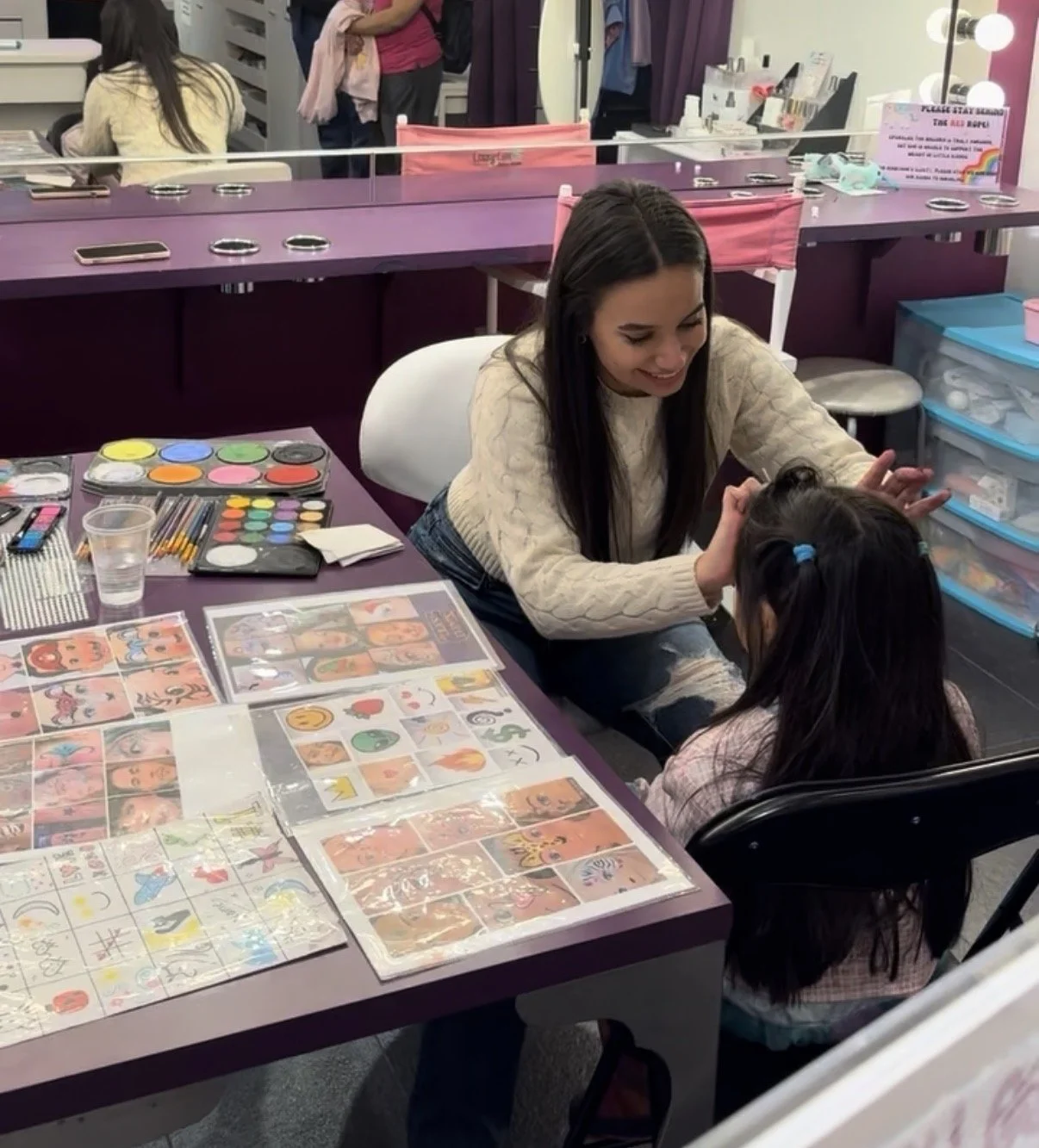 A woman and young girl sitting at a purple table filled with art supplies, stickers, and colorful artwork in a craft room or classroom. The woman is smiling and engaging with the girl, who has her hair tied in pigtails with blue hairbands.