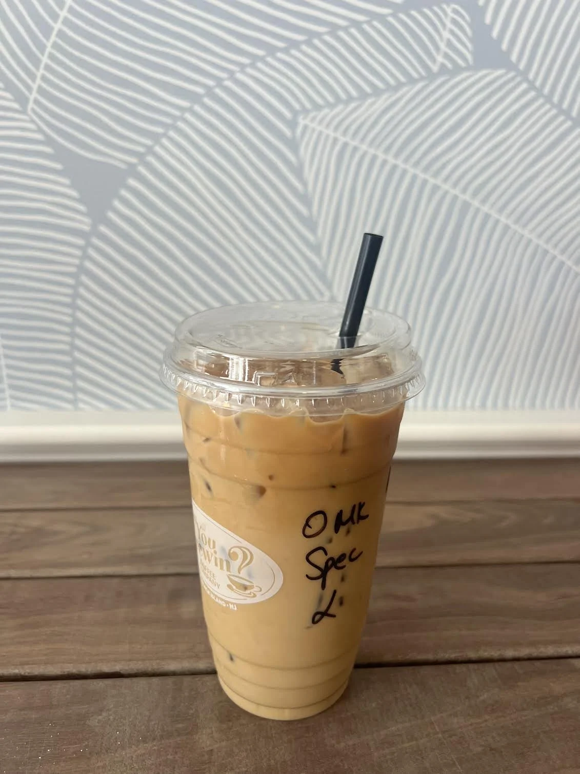 A plastic cup filled with iced coffee topped with a plastic lid and black straw, sitting on a wooden table with a blue patterned wall in the background. Handwritten on the cup is 'O M G Spec L.'