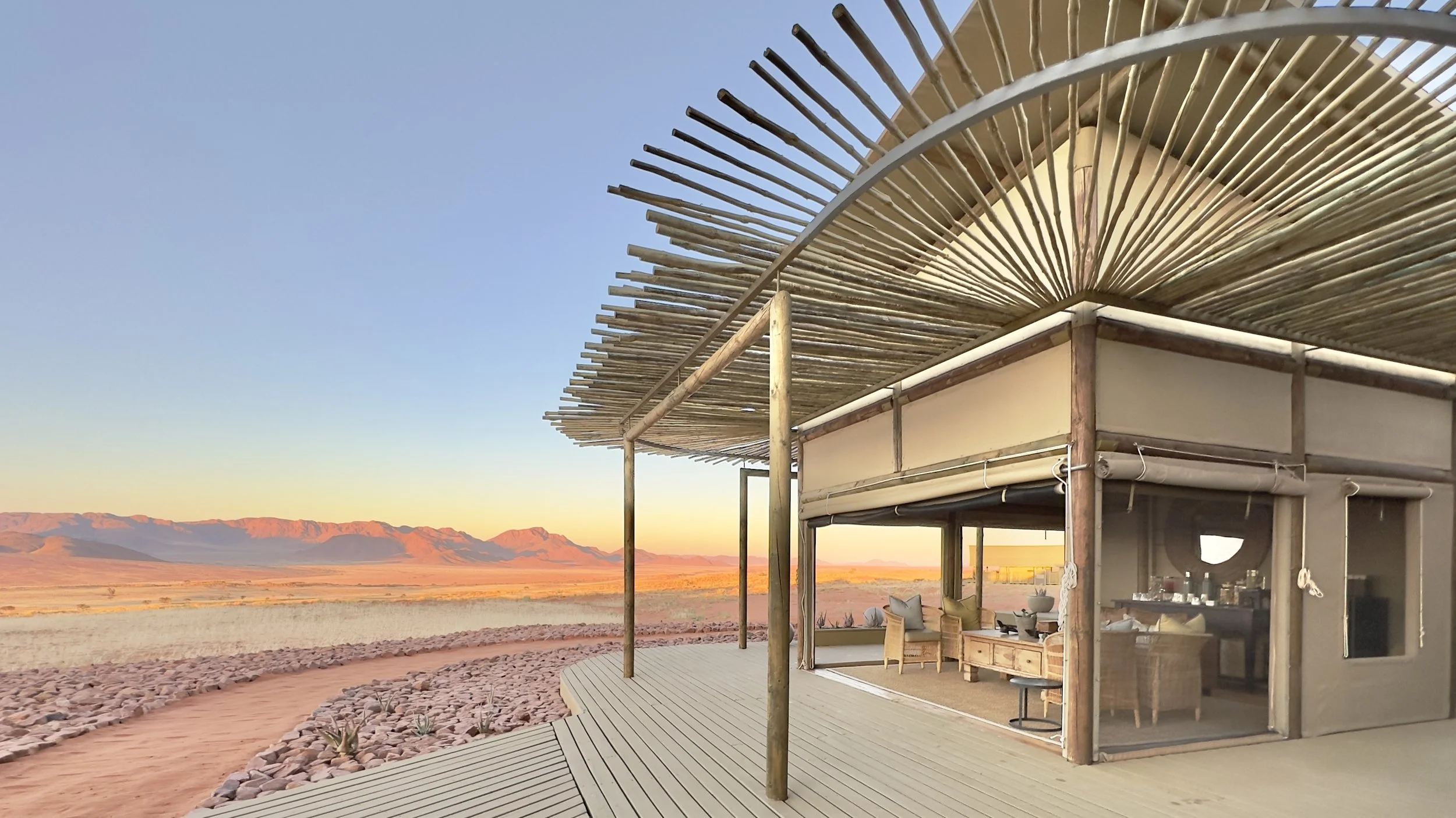 A modern, open-air structure with a bamboo roof in a desert landscape at sunset, mountains in the distance.