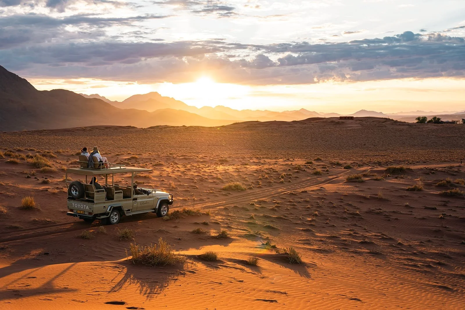 A safari vehicle with an open roof carrying tourists is driving through a desert at sunset, with mountains in the background and scattered bushes on the sand.
