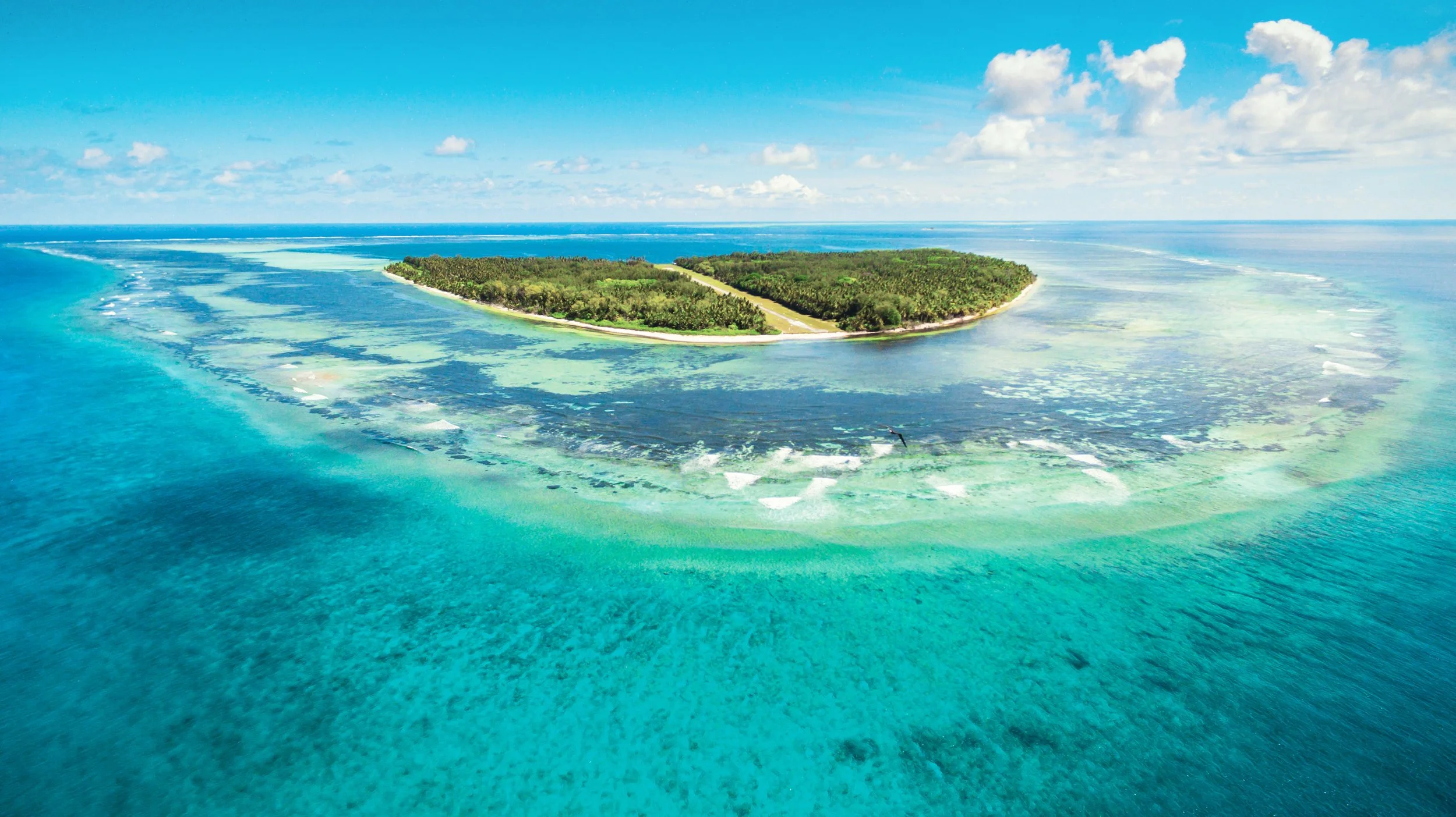 Aerial view of a tropical island with lush green trees, surrounded by turquoise coral reefs and clear blue ocean waters under a partly cloudy sky.