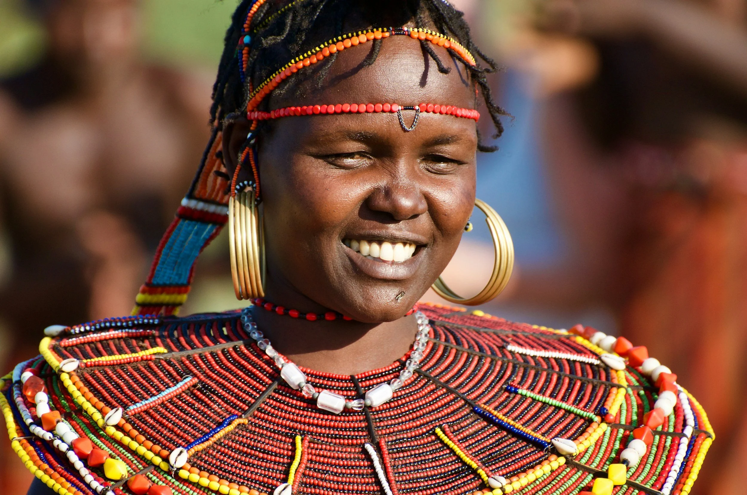 A smiling African woman wearing traditional beaded jewelry and headdress, with blurred background.