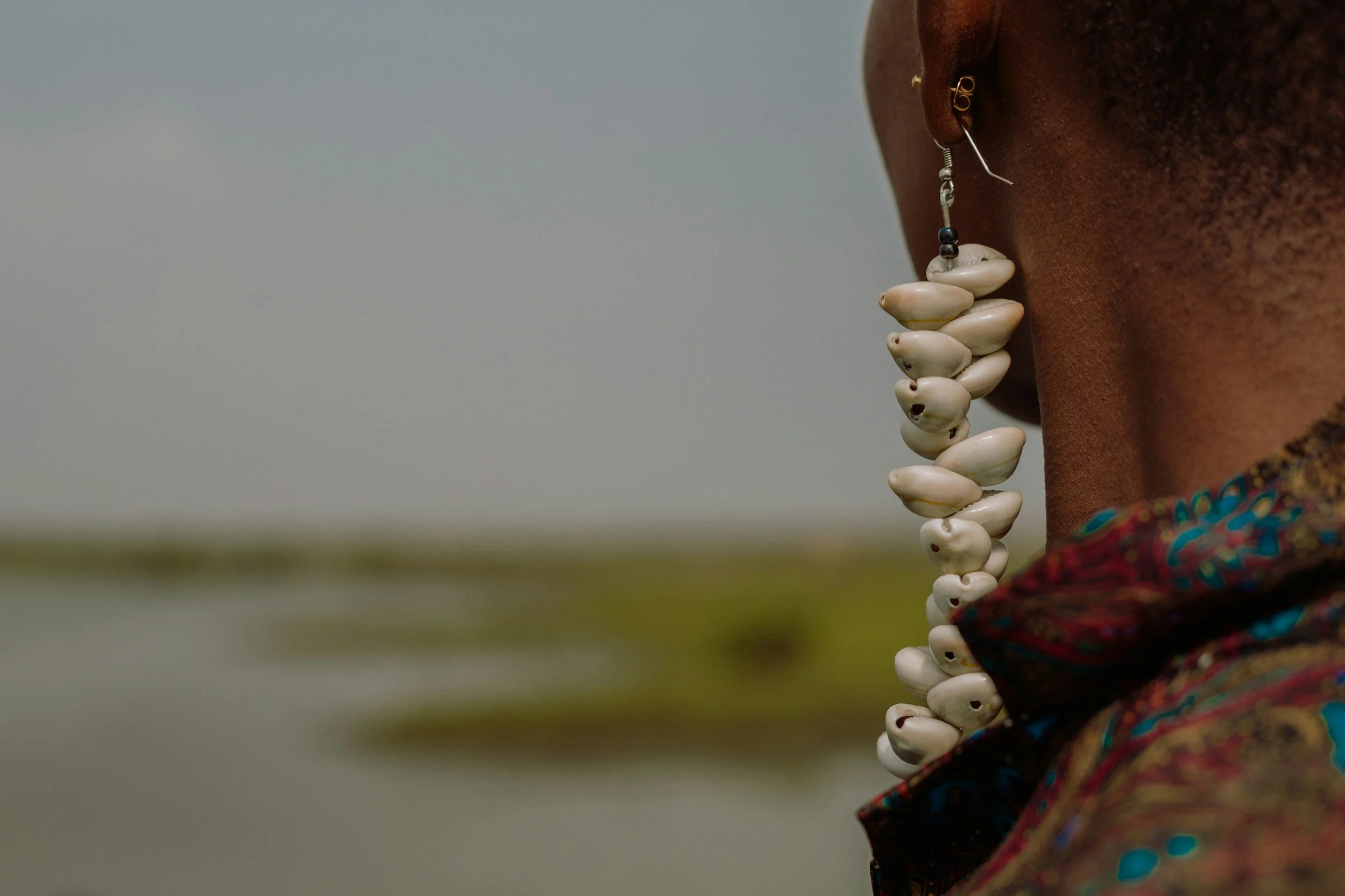 Close-up of a person's side face showing multiple white shells as jewelry on the ear and neck, with colorful fabric on the shoulder, against a blurred outdoor background.