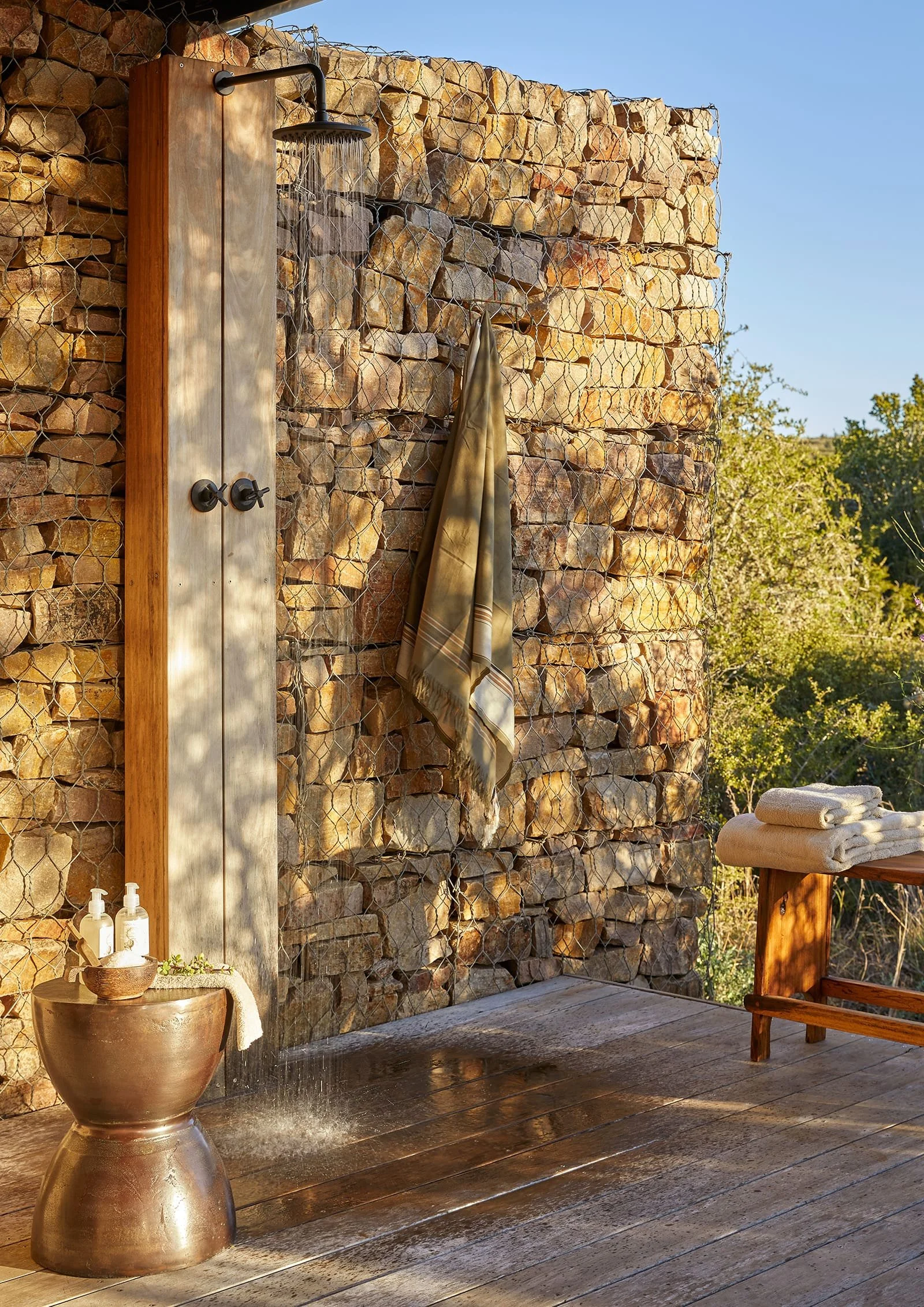 Outdoor shower and seating area with a stone wall, towel, and folded towels on a wooden table.