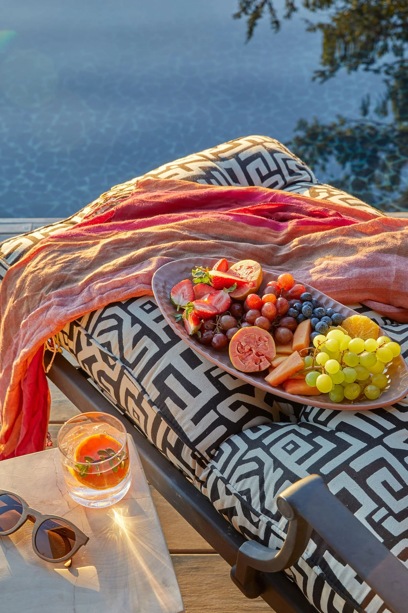 Outdoor scene with a person relaxing by a swimming pool, with a tray of fresh fruit including strawberries, blueberries, grapes, and sliced tomatoes, a glass of fruit-infused water, sunglasses on a table, and a patterned cushion on a lounge chair, du
