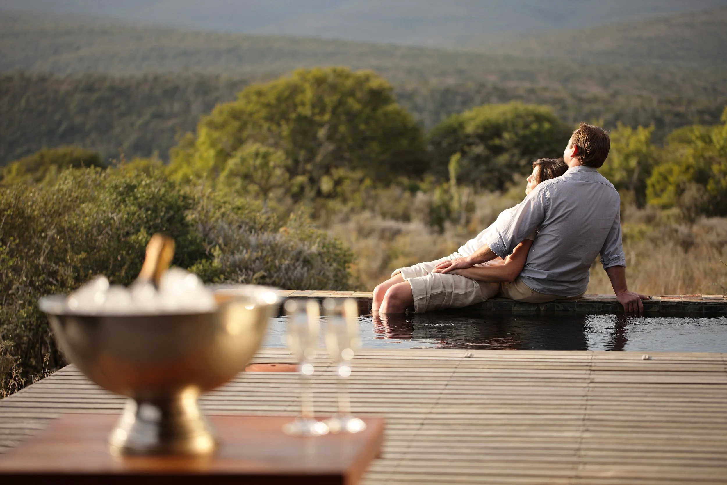 A man and woman sitting by a pool overlooking a hilly landscape with green trees, enjoying a relaxing moment.