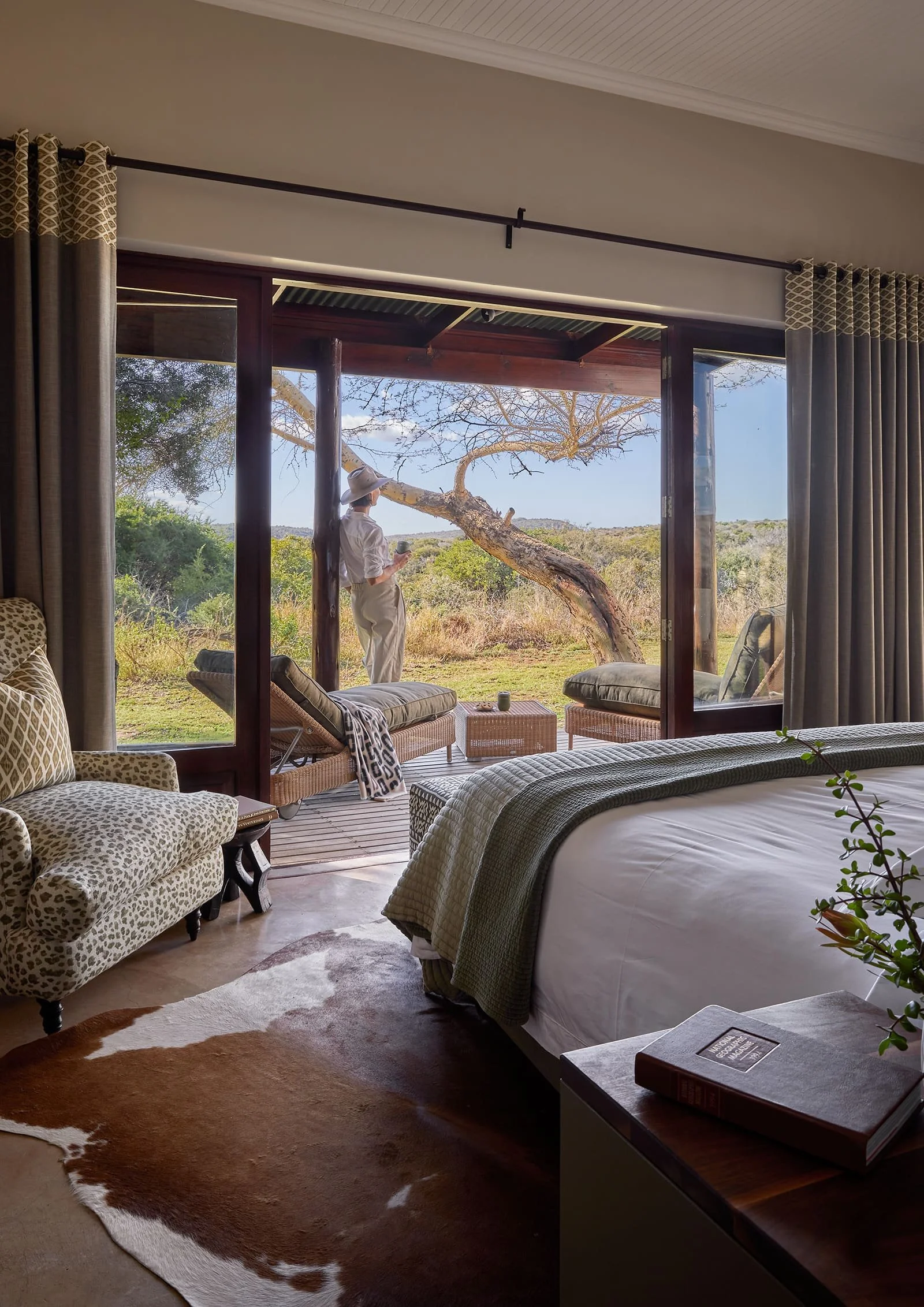 View of a bedroom with an open sliding glass door leading to a porch with a woman in a white hat and clothes standing outside near a tree. Inside the room, there is a bed, a cowhide rug on the floor, a patterned armchair, and a side table with books.
