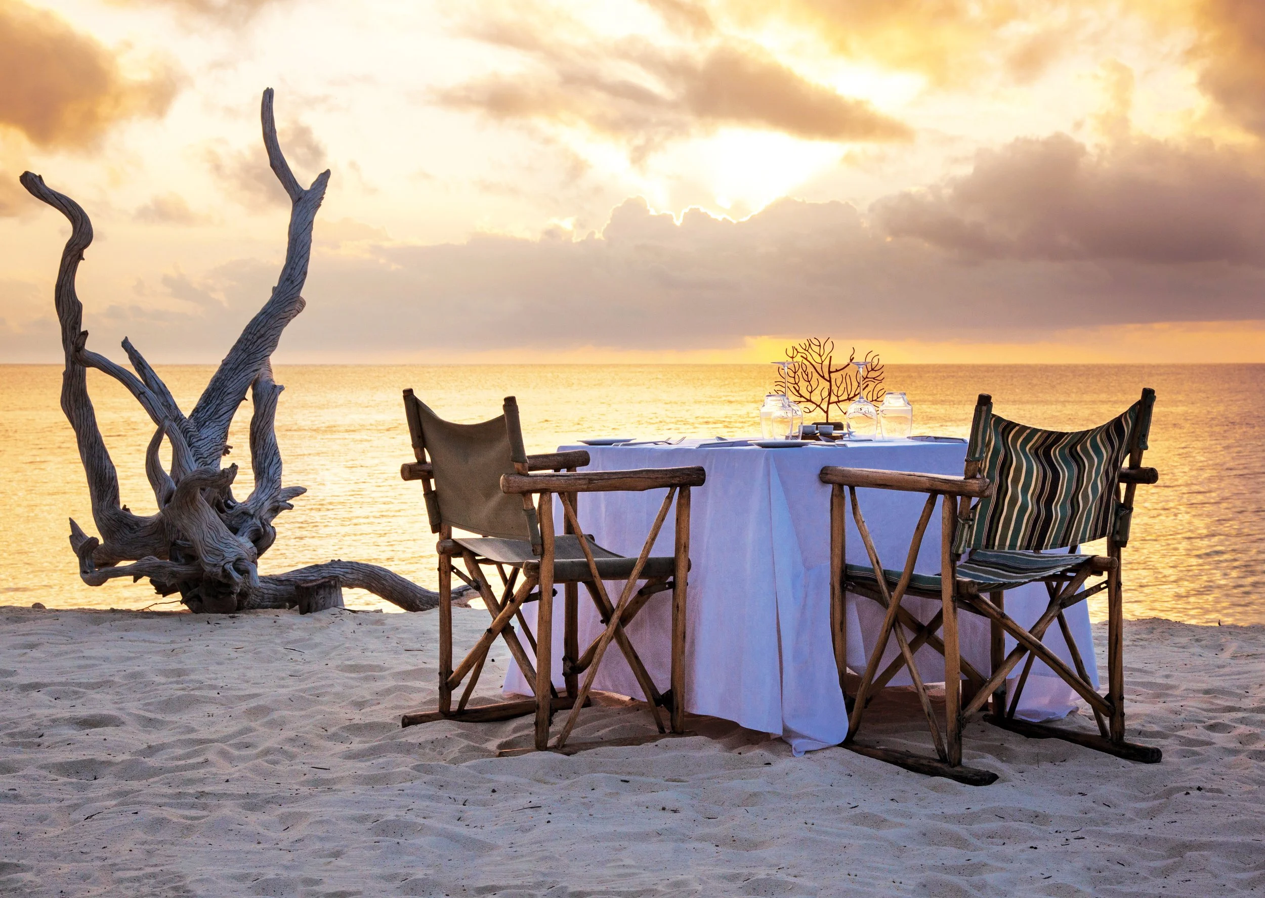 A table set for dining on a beach at sunset, with chairs, water glasses, and a decorative piece, with a driftwood sculpture in the foreground and ocean and cloudy sky in the background.
