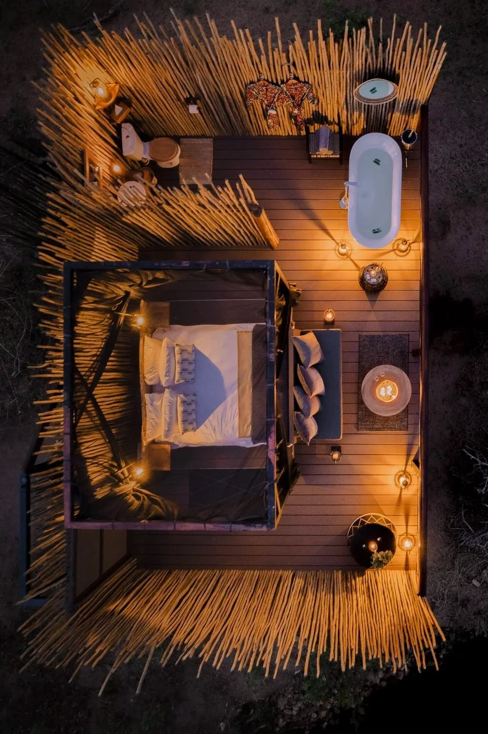 A cozy outdoor bedroom with a bed, a privacy bamboo fence, and string lights. There's a bathtub, a sitting area with a sofa and a coffee table, and decorative lighting creating a warm ambiance.
